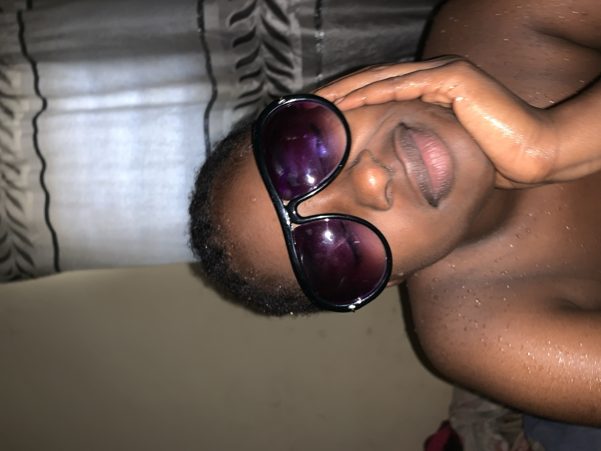 A close-up, portrait-style image depicts a person with dark skin, wearing sunglasses with purple lenses. The person's right hand is touching their forehead. The person's skin appears moist or sweaty. The background includes a wall with a patterned curtain. The location is speculated to be Rafin Par, Nigeria.