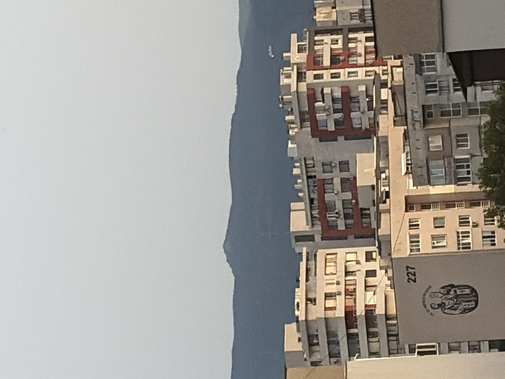 In a medium-shot, the image captures a cityscape with several multi-story apartment buildings in the foreground. The buildings exhibit a mix of architectural styles and colors, with some featuring white and light-colored facades, while others have accents of red. Many windows are visible on the buildings, some with balconies. One building has the number "227" painted on its side, along with a circular image depicting two figures in traditional attire. In the background, a dark, hazy mountain range forms the horizon under a pale sky. A faint white object, possibly a small aircraft or drone, is visible in the sky above the mountains. The overall scene suggests an urban environment with a natural backdrop, likely taken during daylight hours.