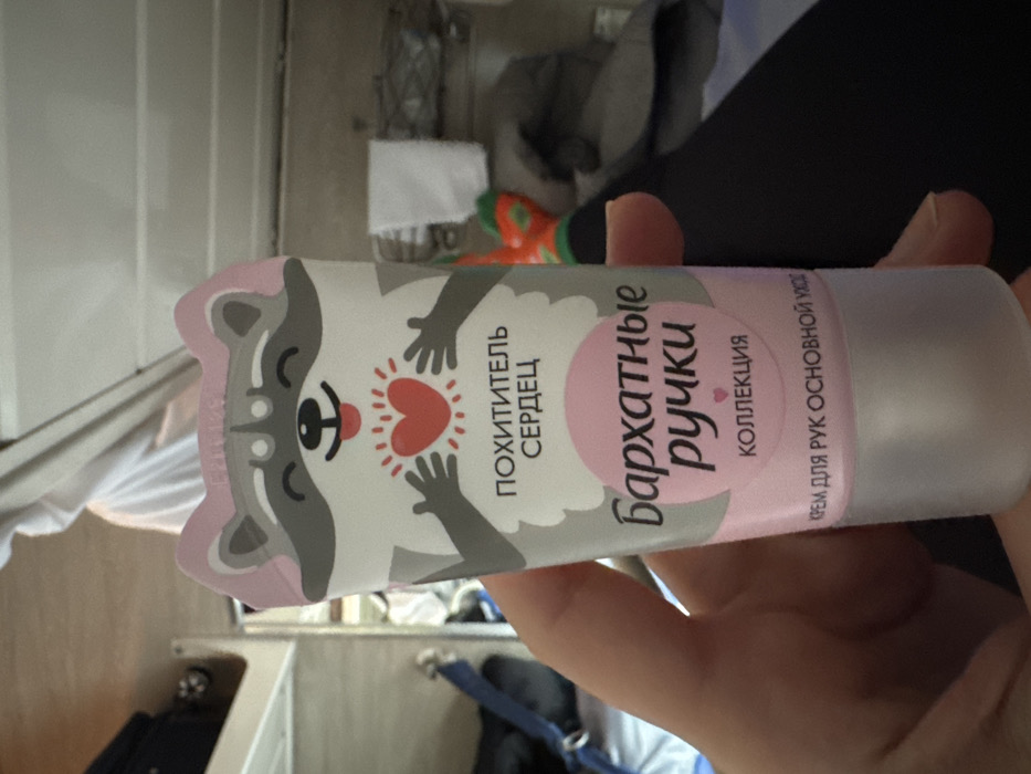 A close-up, slightly upward-angled image depicts a person's left hand holding a tube of hand cream within a train compartment. The hand cream tube is predominantly white and pink, featuring a cartoon raccoon character at the top, depicted with a happy expression and holding a glowing red heart. Russian text on the tube includes "ПОХИТИТЕЛЬ СЕРДЕЦ" (Heart Thief), "Бархатные ручки" (Velvet Hands), "КОЛЛЕКЦИЯ" (Collection), and "КРЕМ ДЛЯ РУК ОСНОВНОЙ УХОД" (Hand Cream Basic Care). The hand is fair-skinned, with visible fingernails, gripping the lower portion of the tube.
The background indicates an interior train setting. To the left, a metallic frame consistent with a bunk bed structure is visible, alongside a blue strap. To the right, a person's lower leg is present, clad in dark trousers and a brightly colored orange sock with green patterns. A white, folded towel hangs on a fixture in the upper right. Other blurred fabric items are discernible in the background. The interior walls and ceiling are light-colored. This scene is set within a train compartment, consistent with travel in Russia, potentially in the vicinity of Gryazi.