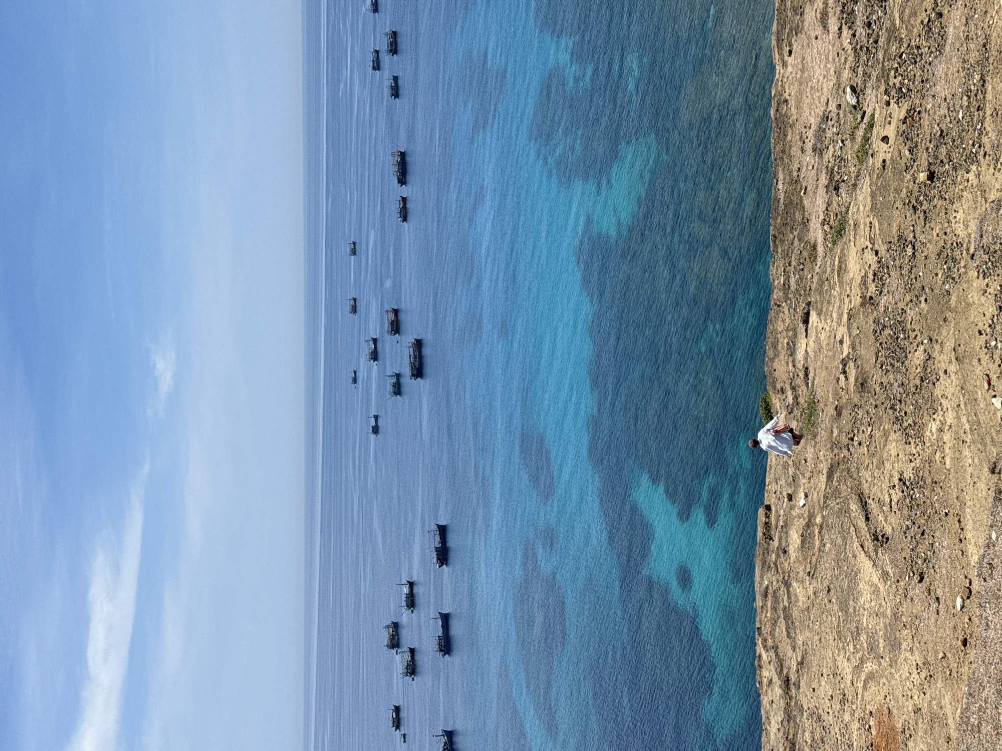 A person walks along a rocky cliff overlooking a bright blue ocean dotted with numerous fishing boats. The person, seen from behind, wears a white shirt and shorts and carries a bag over their shoulder. The water exhibits clear turquoise patches revealing submerged coral reefs beneath the surface. The sky is a pale blue with scattered wispy clouds. The scene depicts a coastal landscape likely in the Lý Sơn Special Administrative Region of Vietnam, known for its islands and fishing activities.