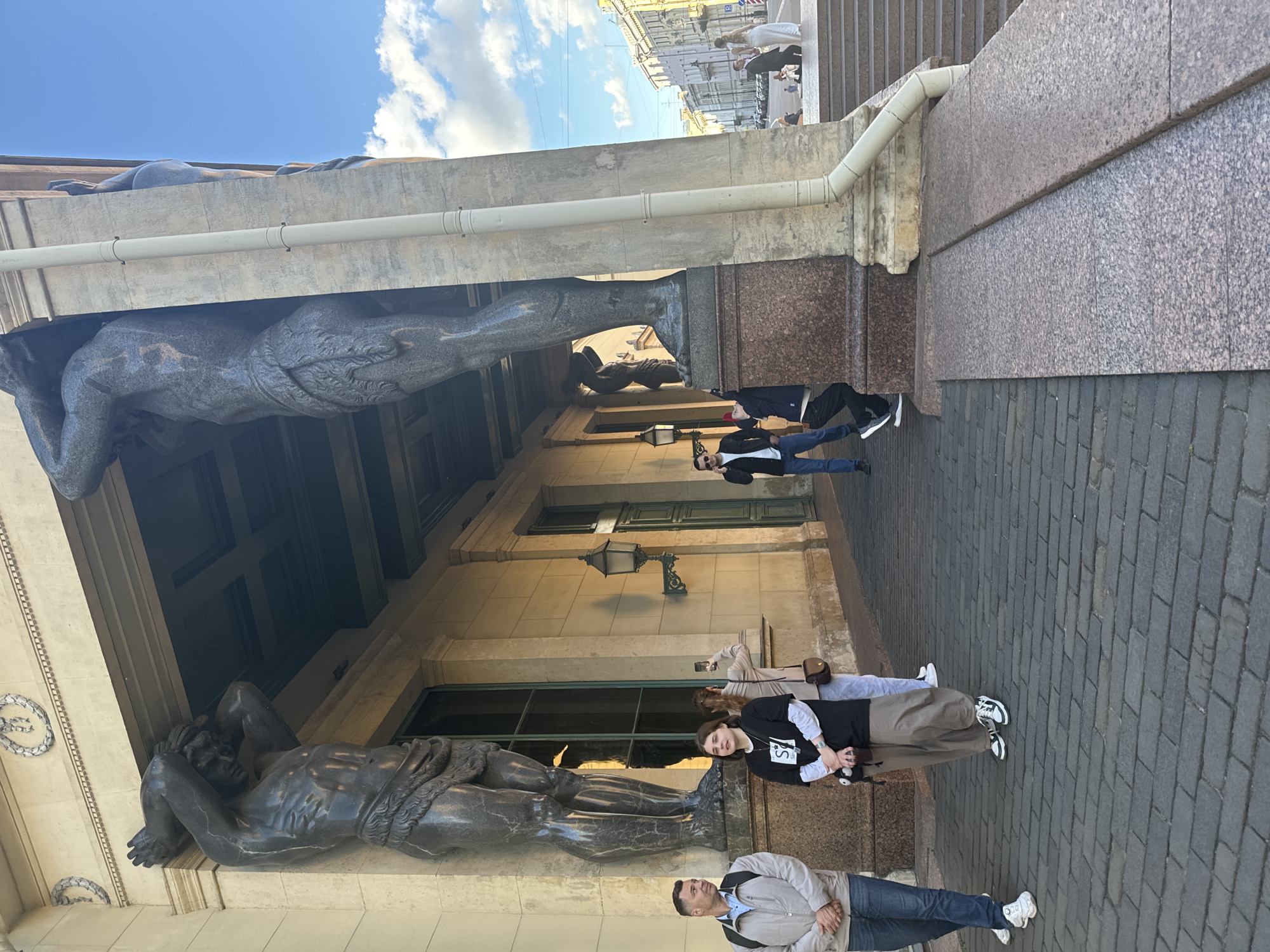 The image depicts the facade of a classical building in Saint Petersburg, Russia, featuring large, dark statues of men holding up the entablature. The building's exterior is a light-colored stone, with tall windows and decorative elements. In the foreground, several people are visible. On the left, a man in a light gray jacket and blue jeans stands with his hands clasped. Next to him, a young woman in a black t-shirt with white graphics and light brown pants is looking at the camera. Further back, another woman is taking a picture with her phone. A man in a dark suit and white shirt is walking in the center of the frame. On the right side of the image, a set of stone stairs leads up to a higher level, and in the distance, a wedding couple is posing for a photo. The sky is blue with scattered white clouds. The ground in front of the building is paved with dark cobblestones. The overall scene suggests a public space, likely a tourist attraction, in the historic center of the city.