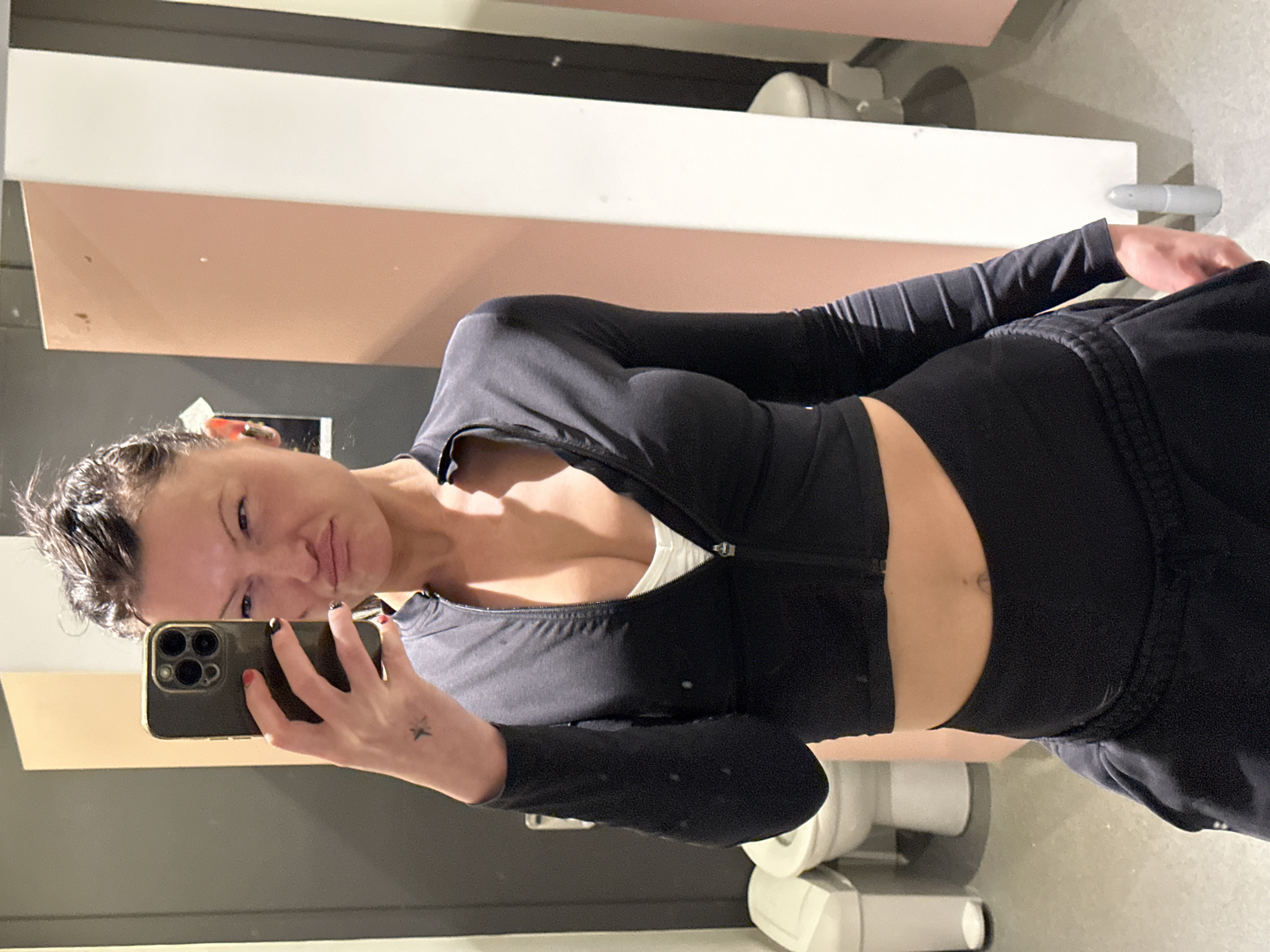 A female subject is depicted in a mirror selfie taken within a public restroom in Tunbridge Wells, United Kingdom. She stands centrally, capturing her reflection with a black smartphone featuring a gold-colored case, held in her right hand. Her dark hair is slicked back and appears damp. Her facial expression features pursed lips and slightly narrowed eyes. She is dressed in a black, long-sleeved, cropped athletic top with a partially unzipped front revealing a white undergarment, and black athletic trousers. A small star tattoo is visible on her right forearm, and her right index fingernail is painted red while others are black. A gold earbud is present in her right ear. The background consists of multiple public restroom stalls, indicated by at least two white ceramic toilets partially visible behind pale pink and white partition walls. A tall, white, lidded waste bin is positioned on the floor to the left. The floor is light grey. A small, grey, cylindrical object rests on the floor near a white partition on the right.