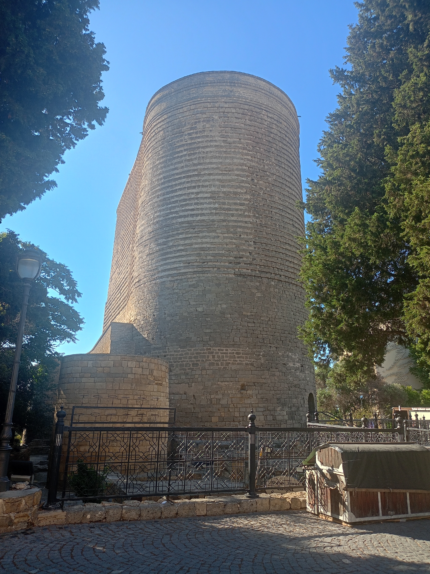 An eye-level shot shows the Maiden Tower in Baku, Azerbaijan. The cylindrical stone tower, constructed from light-colored bricks arranged in horizontal bands, dominates the frame. A curved, lower stone structure abuts the base of the tower on the left. In the foreground, a black metal fence with intricate geometric patterns encloses the area around the tower. A cobblestone pathway covers the ground. To the right of the fence, a small, wooden structure with a dark canvas roof sits on the cobblestones. Green trees frame the tower on the left and right, and a clear blue sky is visible above. A lamppost stands to the far left of the frame. The scene is illuminated by bright sunlight, casting shadows on the ground.
