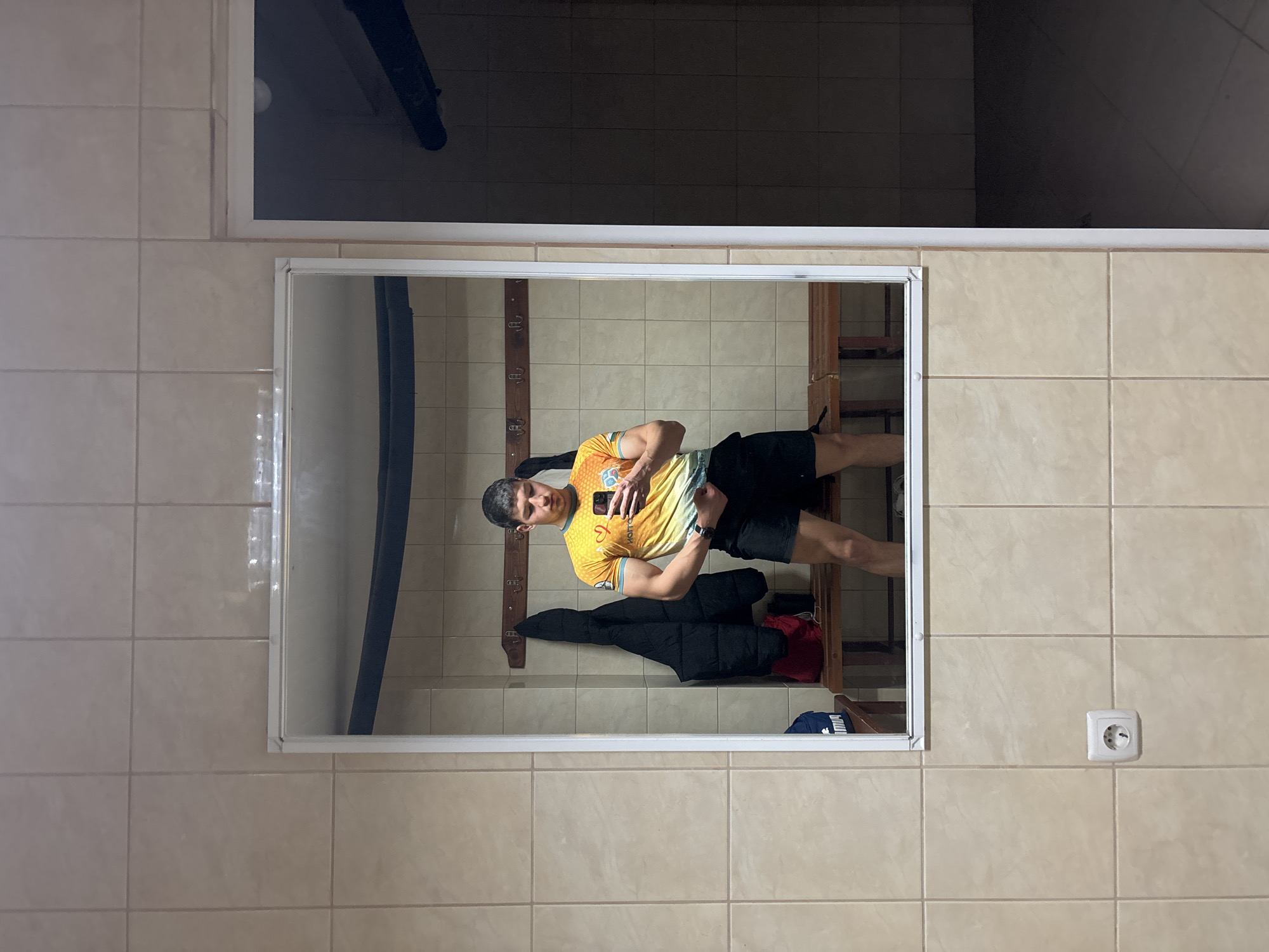 A man is taking a selfie in a mirror. He is wearing a yellow and orange soccer jersey and black shorts. The mirror is located on a wall covered in beige tiles. Above the mirror is a dark rectangular area, possibly another mirror or a window. To the right of the mirror is a white electrical outlet. The reflection in the mirror shows a coat rack with a jacket hanging on it, and a shelf with various items, including clothing. The scene appears to be a changing room or bathroom. This picture was taken in Zlatograd, Bulgaria.