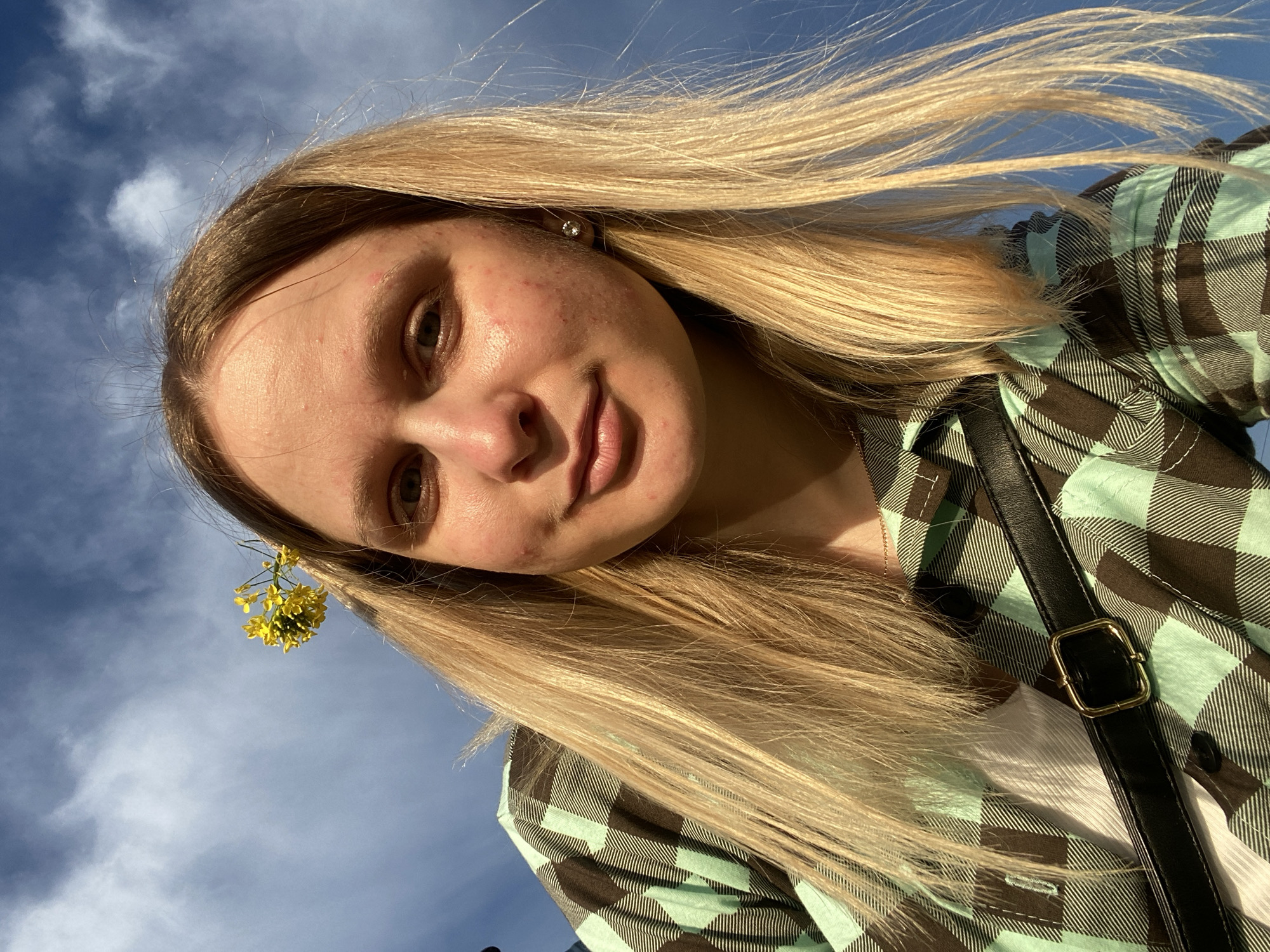 A young woman is captured in a selfie, looking directly at the camera with a subtle, pleasant smile. Her face is slightly tilted, and her long, straight blonde hair appears windswept, catching the warm natural light. Her eyes are light-colored, and some minor skin blemishes are visible, suggesting a natural and unedited appearance.
She is dressed in a plaid shirt featuring a pattern of dark brown and light blue/mint green squares, worn over a white garment. A black strap with a gold buckle, likely part of a bag, is visible across her chest. A small, sparkling stud earring is worn in her right ear, and a cluster of bright yellow flowers is tucked into her hair near her left ear, adding a decorative touch.
The photograph is taken from a low angle, with the vibrant blue sky and scattered white, wispy clouds forming the background. The strong natural light indicates it is daytime, likely in the afternoon. The scene takes place outdoors in Chaikovsky, Russia. No text is visible in the image.