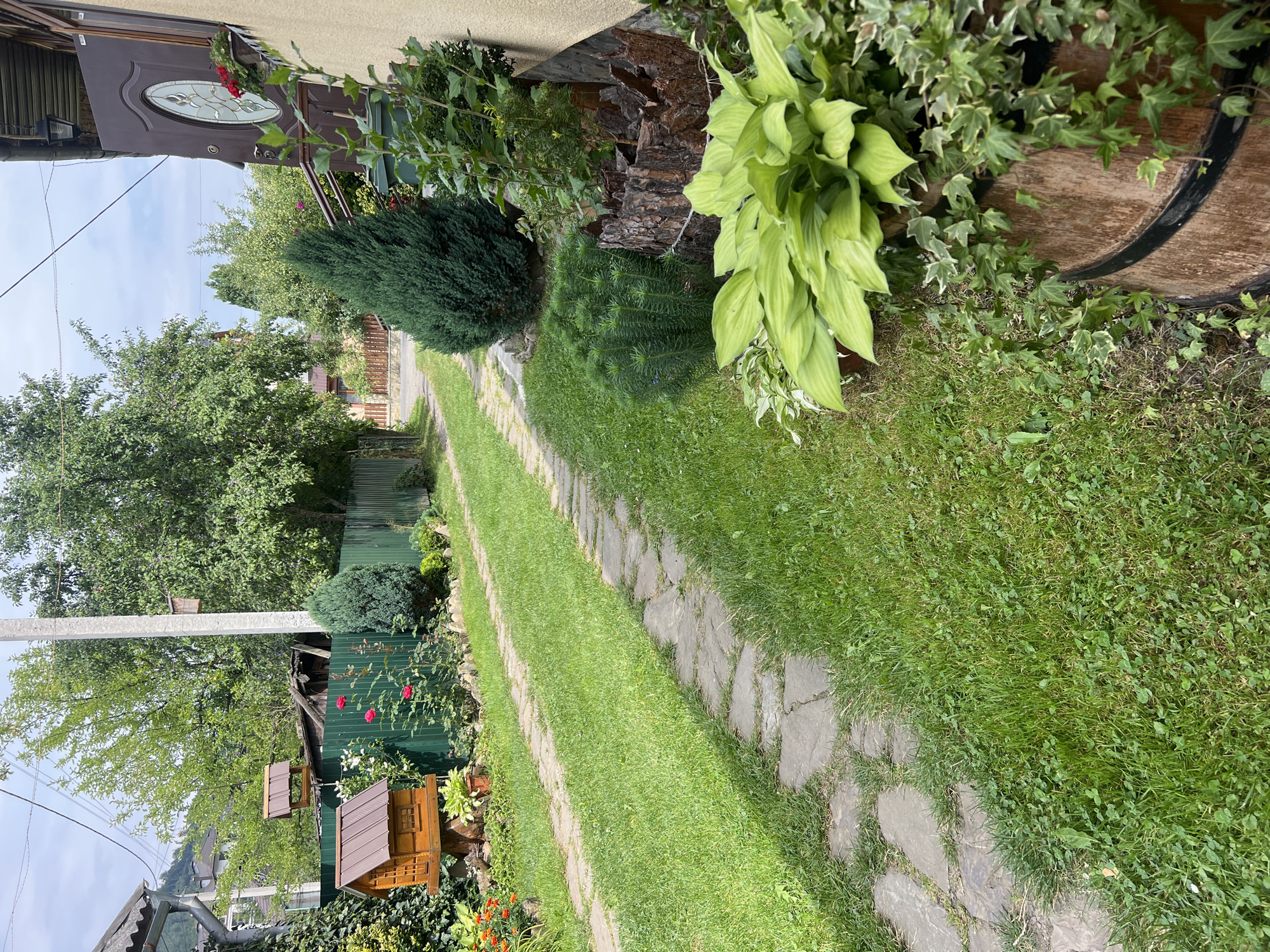 The image depicts a tranquil, green pathway in what appears to be a residential garden. A stone-paved path meanders through a well-maintained lawn, bordered by lush greenery and flowering plants. To the left, a wooden birdhouse stands prominently, suggesting a connection to nature. The pathway leads towards a residential building, partially visible on the right, with a characteristic dark brown door and a window. The surrounding environment includes trees, a fence, and hints of distant buildings, possibly indicating a suburban or semi-rural setting. The overall impression is one of a peaceful, natural setting within a populated area, possibly a backyard in Yaremche, Ukraine, given the architectural and horticultural context often found in the region.