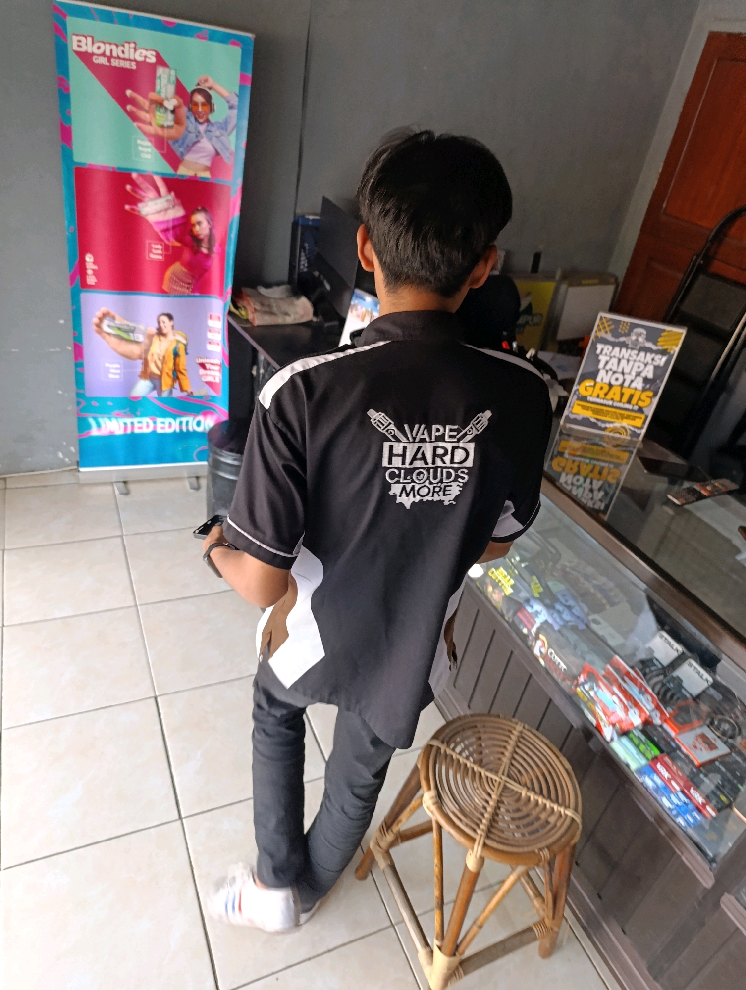 The image depicts the rear view of a young man standing in what appears to be a small retail shop or stall in Karangsono, Indonesia. He is wearing a black polo shirt with white and brown accents, and the back of the shirt features white text that reads "VAPE HARD CLOUDS MORE" along with two stylized images of vape devices. He is also wearing dark pants and white sneakers. The man is holding a smartphone in his right hand and looking down.
In the background, to the left, is a tall, colorful promotional banner for "Blondies Girl Series." To the right, there is a glass display counter filled with various vape-related products, such as e-liquids and accessories. A small, circular stool made of woven rattan and wood is positioned in front of the counter. A sign with text in Indonesian, including "TRANSAKSI TANPA NOTA GRATIS," is placed on the counter, indicating a promotion for transactions without a receipt. The floor is tiled with light-colored squares. The overall environment suggests a business that sells vaping products.