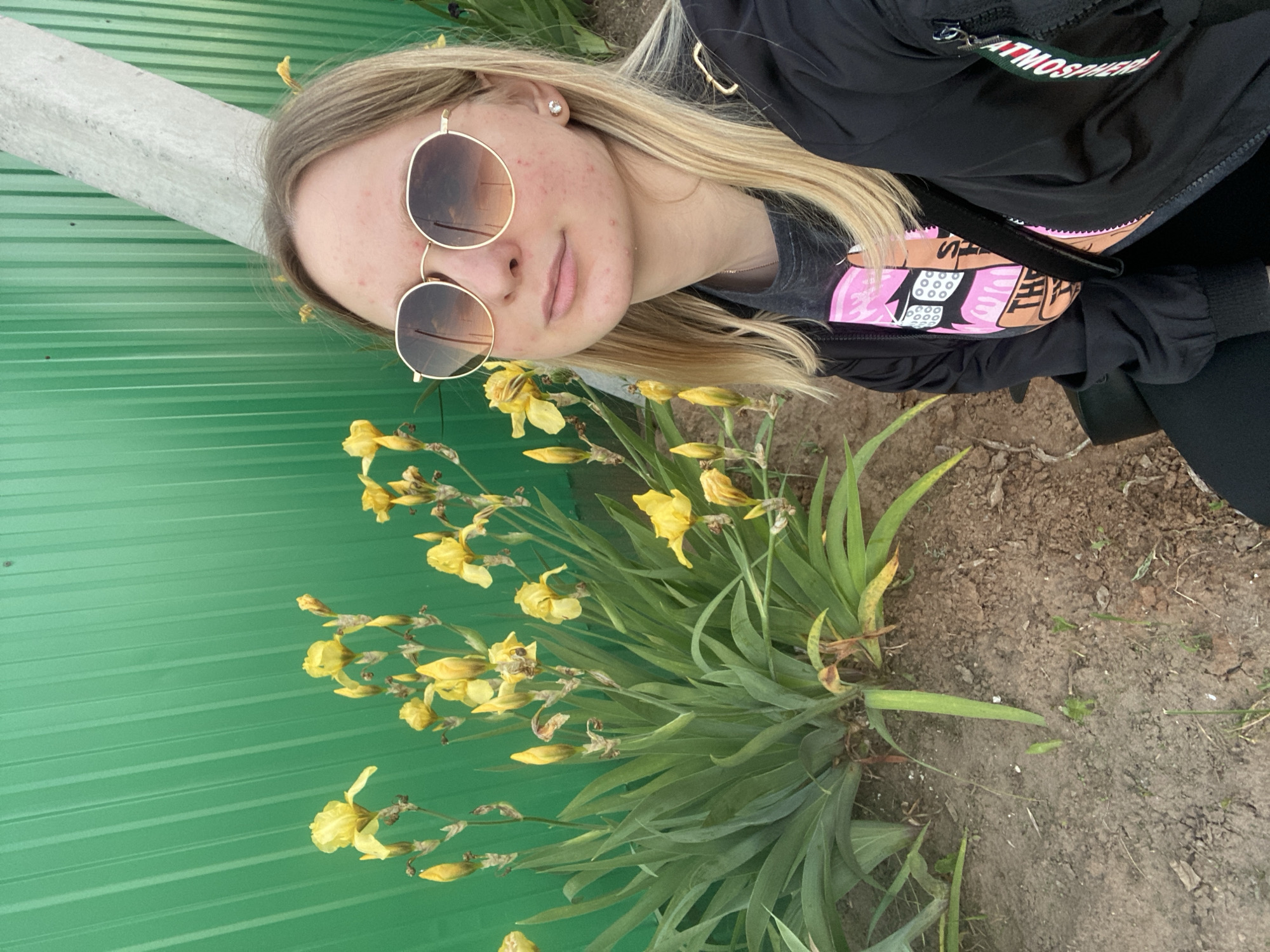 The image displays a self-portrait of a young adult female captured outdoors. She is positioned on the right side of the frame, wearing round, gold-rimmed sunglasses with gradient brown-orange mirrored lenses reflecting a cloudy, potentially sunset-lit sky. Her facial skin exhibits multiple scattered red blemishes, and she has long blonde hair. A small, clear stud earring is visible in her right ear. She is dressed in a black jacket and a dark-colored t-shirt featuring a graphic design with pink elements and partial text reading "THE". A black strap crosses her chest.
To the left and behind the subject is a dense cluster of yellow iris flowers and buds, appearing in various stages of bloom and decay. These flowers occupy a significant portion of the midground and left foreground. The background is a bright green corrugated metal fence or wall, running horizontally. A thick, light-colored diagonal pole or beam extends from the upper right of the frame, passing behind the subject's head and resting against the green fence. The foreground consists of dry, brown soil with minimal, sparse green vegetation. The location is Unknown, Unknown.