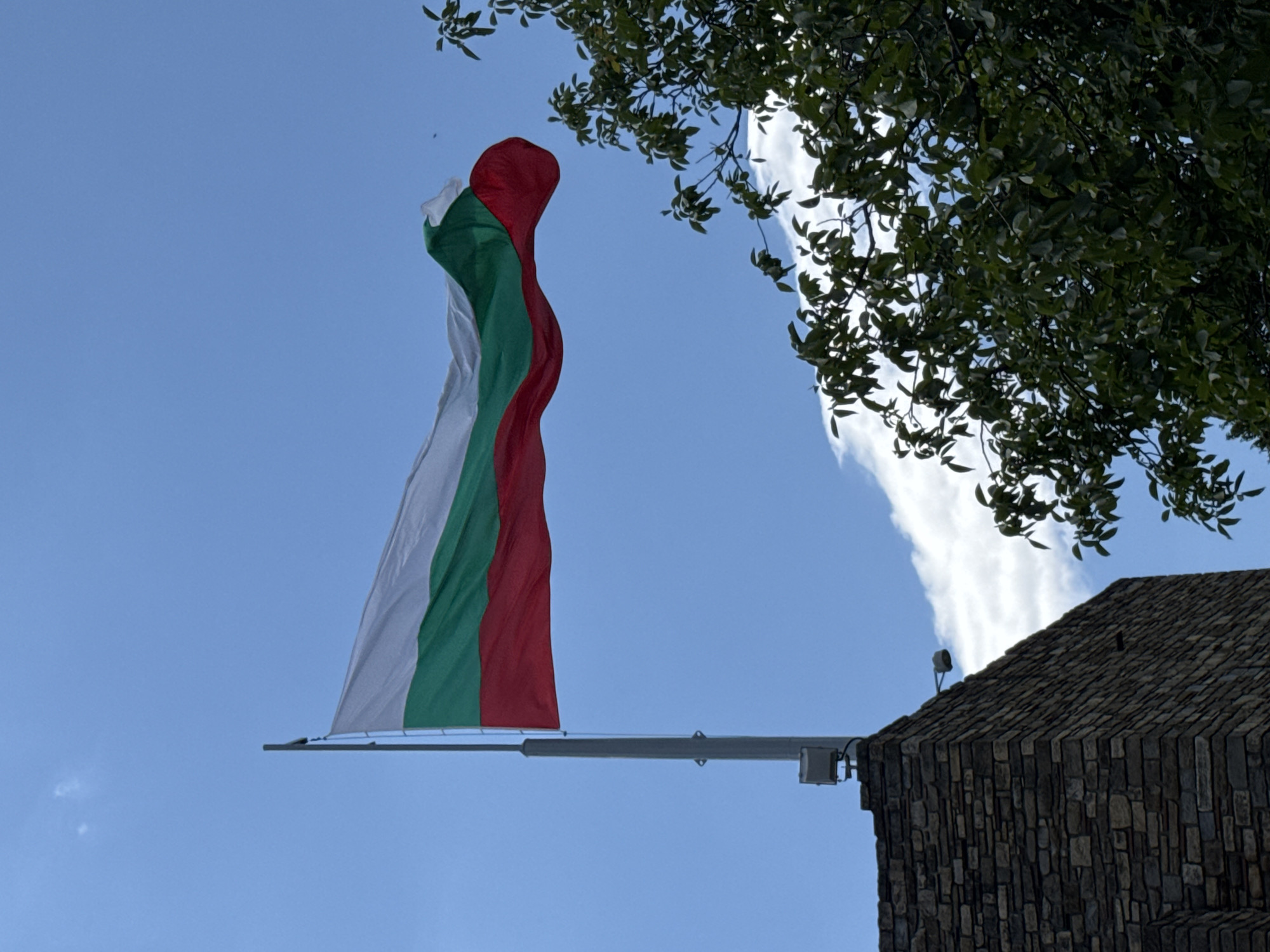 The image shows the Bulgarian flag waving against a blue sky with some clouds. The flag has three horizontal stripes: white on top, green in the middle, and red on the bottom. A flagpole supports the flag, and a stone building is visible in the lower left corner. There is also a tree with green leaves in the lower right portion of the image. The building appears to be a brick structure with a textured surface. There are two light fixtures visible on the top of the building.