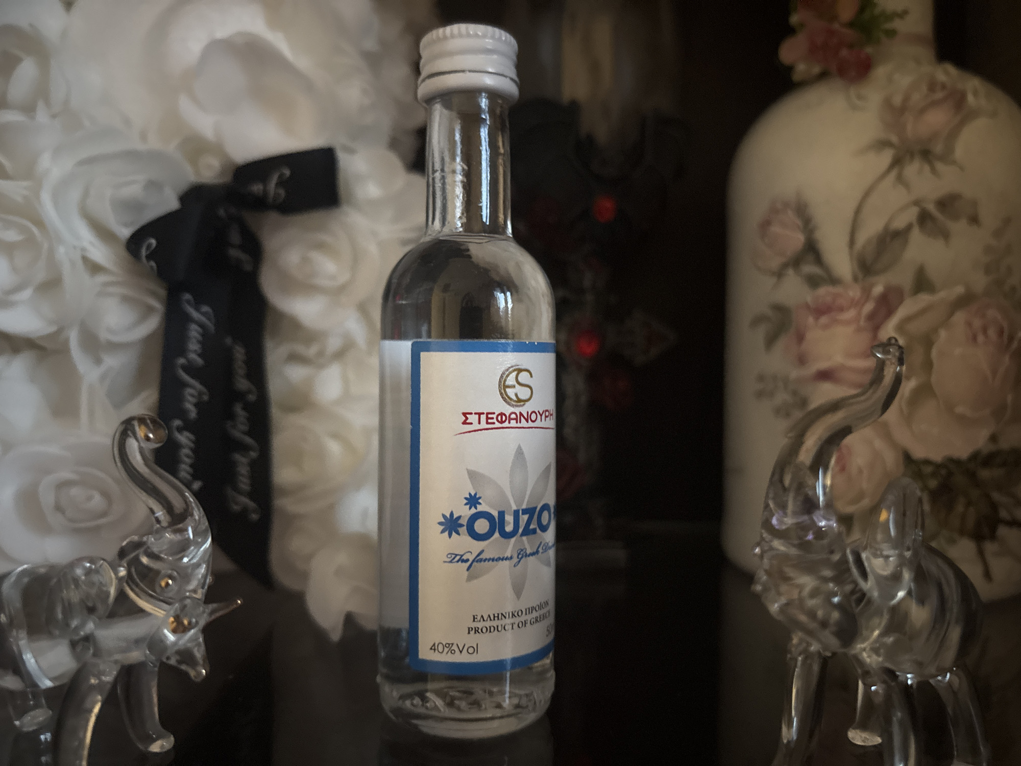 The image shows a miniature bottle of "Ouzo" liqueur, with a white label and blue accents. The label indicates the liqueur is a product of Greece. The bottle stands on a dark, reflective surface, possibly a tabletop or shelf. The background is cluttered with decorative objects, including a white floral arrangement tied with a black ribbon that reads "Just for you," a dark, ornate cross, and a floral-patterned vase. Two small, clear glass elephant figurines flank the bottle on either side. The environment appears to be an indoor setting, likely a home interior. There is no indication of this being taken in Plovdiv, Bulgaria.