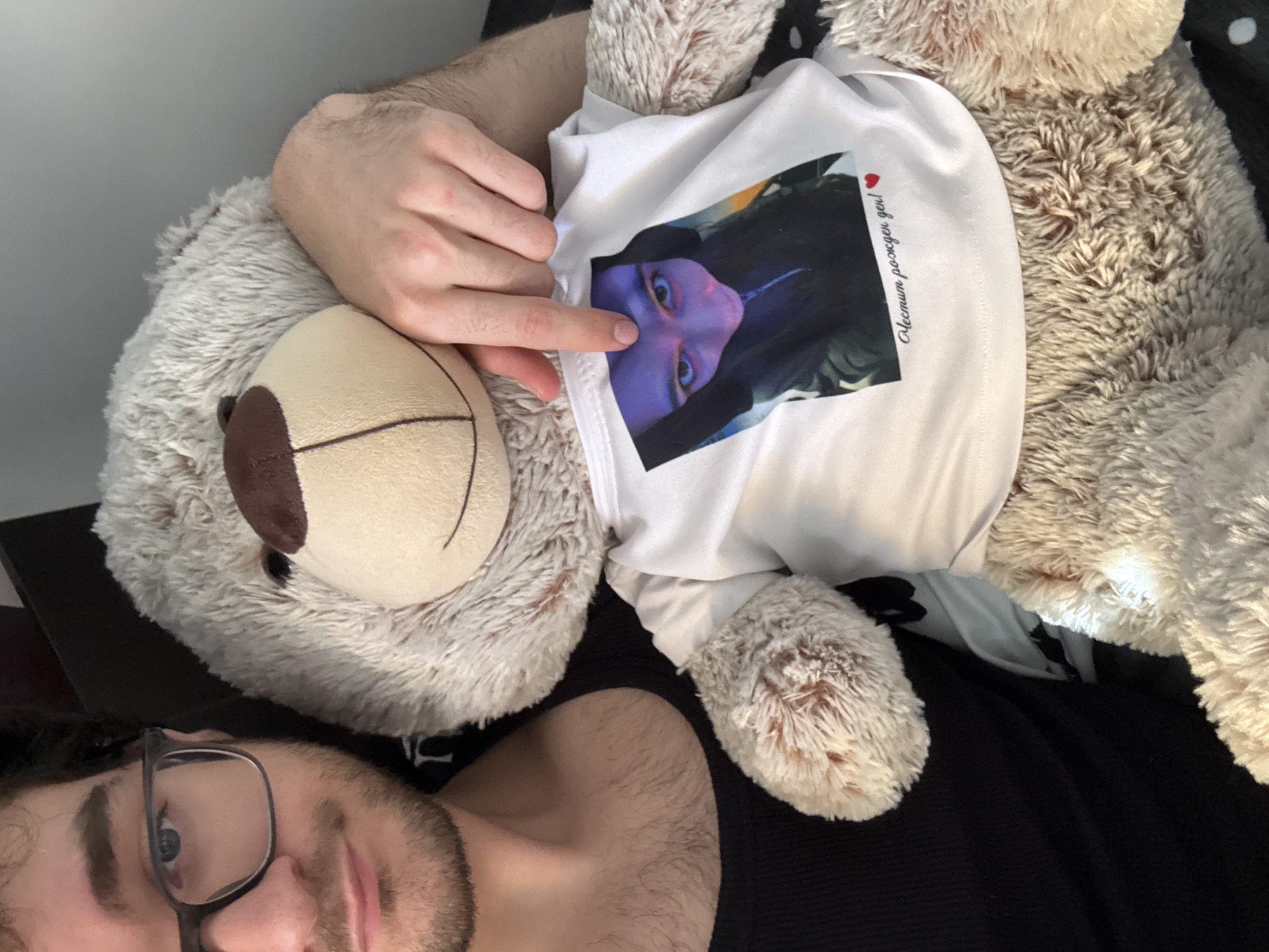 The image captures a man and a large teddy bear lying together, viewed from an overhead perspective. The man is visible in the lower left quadrant, wearing glasses and with visible facial hair (beard/stubble), looking towards the camera. His right arm and hand are draped over the teddy bear. The teddy bear, which is light brown with a darker snout, is wearing a miniature white T-shirt.
On the teddy bear's T-shirt, there is a printed image of a person's face, dramatically lit with blue and purple tones, giving an intense visual effect. The man's index finger is pointing directly at this printed face. To the right of the image on the shirt, text in Cyrillic script is visible, reading "С днем рождения!" (Happy Birthday!), followed by a small red heart icon. The setting appears to be indoors, suggesting a relaxed and personal environment. The image details imply the teddy bear is a personalized gift. The location context for the image is Blagoevgrad, Bulgaria.