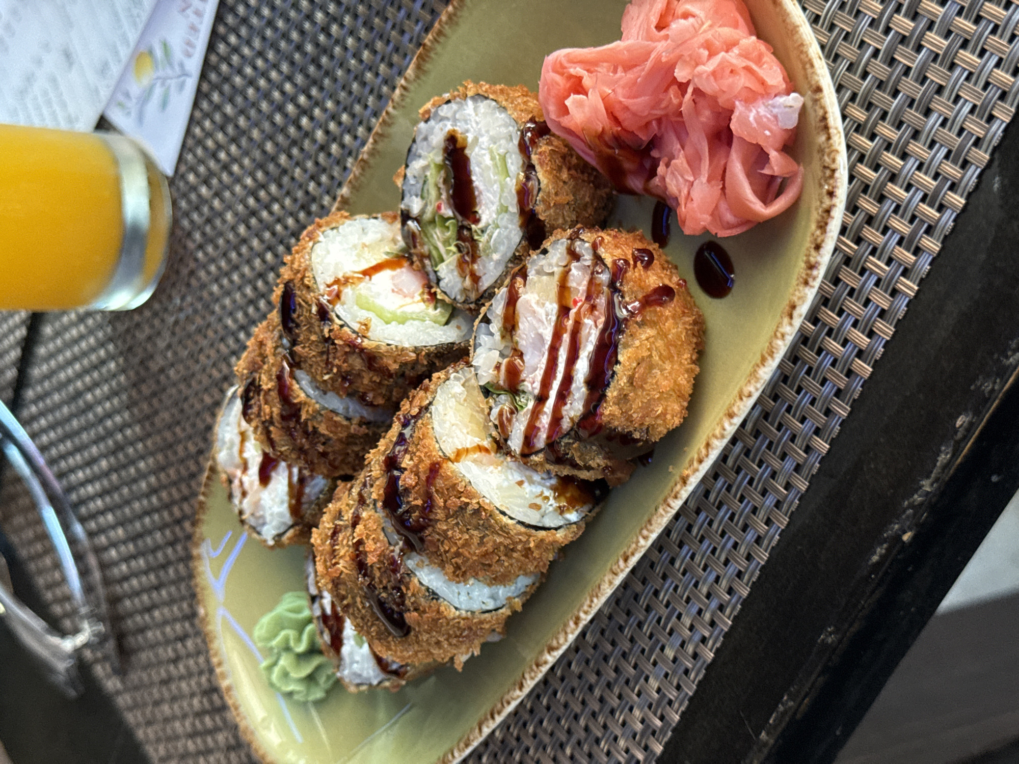 In Budva, Montenegro, a plate of fried sushi rolls is presented on a woven placemat. The plate, a light green with a brown rim, holds six sushi rolls cut in half. Each roll is coated in a crispy panko breading and drizzled with a dark sauce. Beside the sushi, a small mound of bright pink pickled ginger is visible. To the left of the rolls, a dollop of bright green wasabi is present. In the background, a glass of orange juice and a paper napkin with a floral design are partially visible. The scene appears to be set on a table with a dark, reflective surface and a textured placemat.