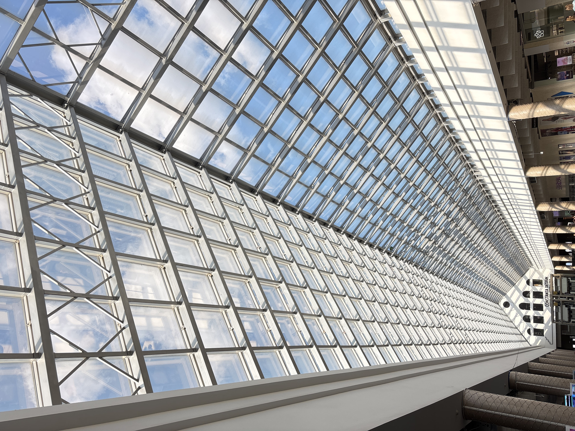 The image captures the interior of a building in Minsk, Belarus, featuring a large glass roof supported by a grid of white metal beams. The sky and clouds are visible through the glass panels. The building's interior shows walls with architectural features, including pillars and upper-level windows. The scene appears to be a public space with commercial establishments visible in the background.