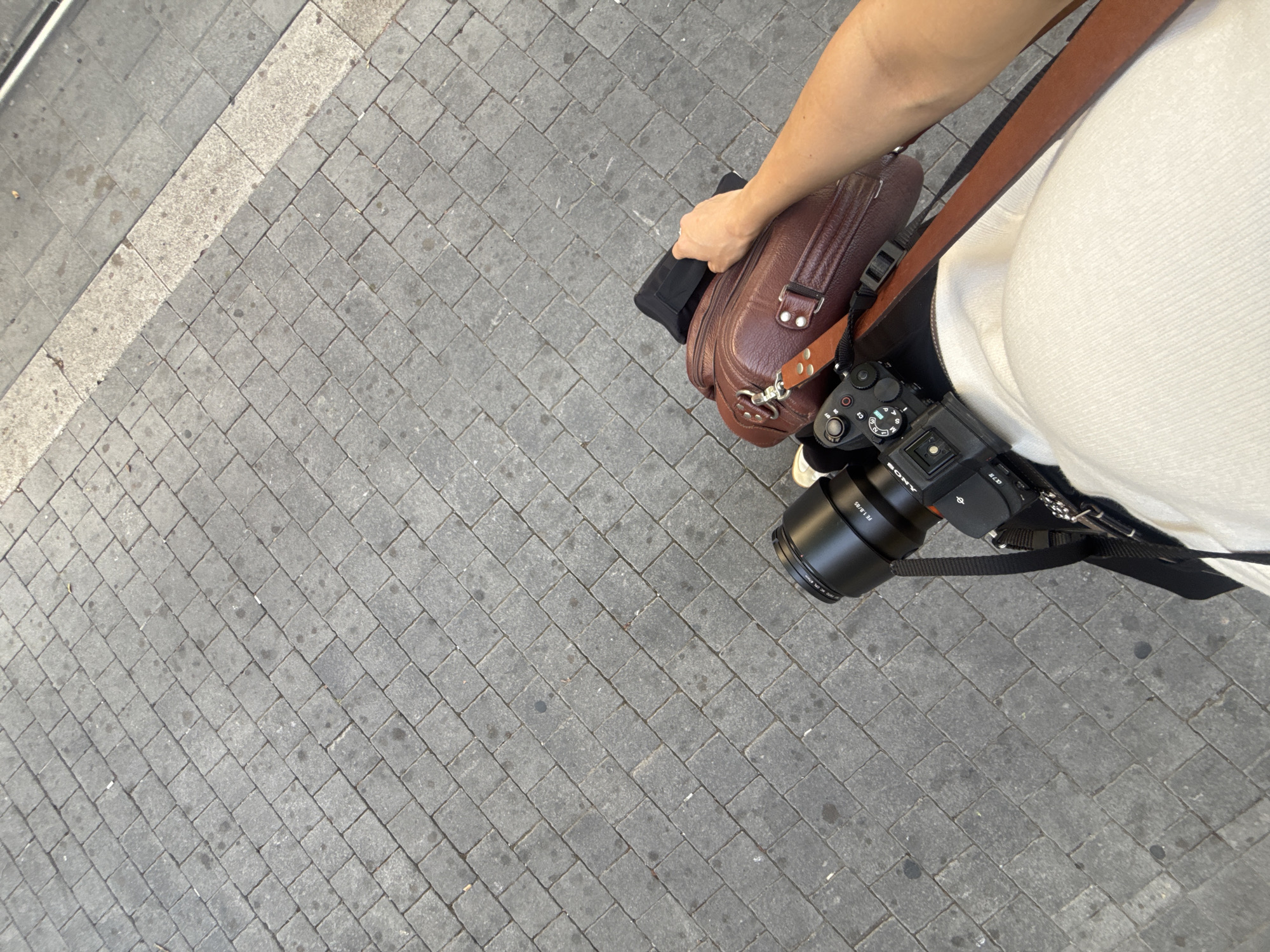 Here's a forensic description of the image:
**Setting:** A paved pedestrian walkway in Tel Aviv, Israel. The surface is composed of grey rectangular cobblestones laid in a herringbone pattern. A lighter, possibly granite, stone border runs along the upper left edge of the frame.
**Subject:** A person is standing on the walkway, seen from a high-angle perspective. The individual is wearing a white top and has a brown leather bag in their left hand. A black Sony camera with a black lens (likely an 85mm based on lens markings) is strapped across their body with black and brown straps.
**Objects:**
* **Leather Bag:** Brown leather, rectangular, vintage-style.
* **Camera:** Black Sony mirrorless camera with attached lens. Lens is inscribed with "FE 1.8/85".
* **Camera Straps:** Black and brown.
**Actions:** The person is standing still or walking slowly. Their left hand is holding the leather bag. The camera is positioned near their waist, ready for use.