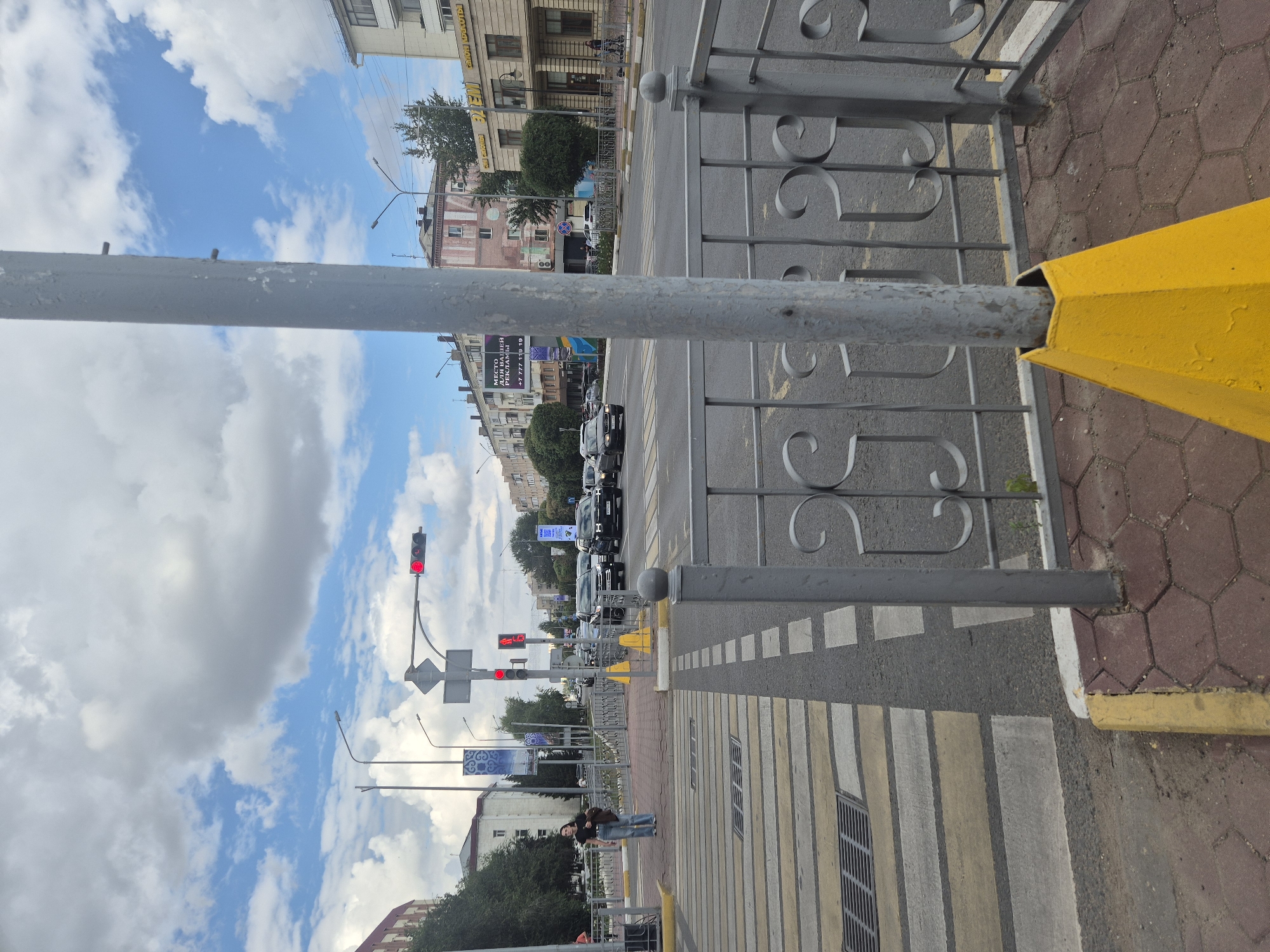 A pedestrian is crossing a street at a crosswalk. The pedestrian is a woman wearing blue jeans and a dark t-shirt, carrying a brown bag. The street is paved and has lane markings. There are traffic lights and street signs at the intersection. Several cars are visible on the road. Buildings line the street, and trees are scattered throughout the area. The sky is cloudy with patches of blue. This image was taken in Kokshetau, Kazakhstan.