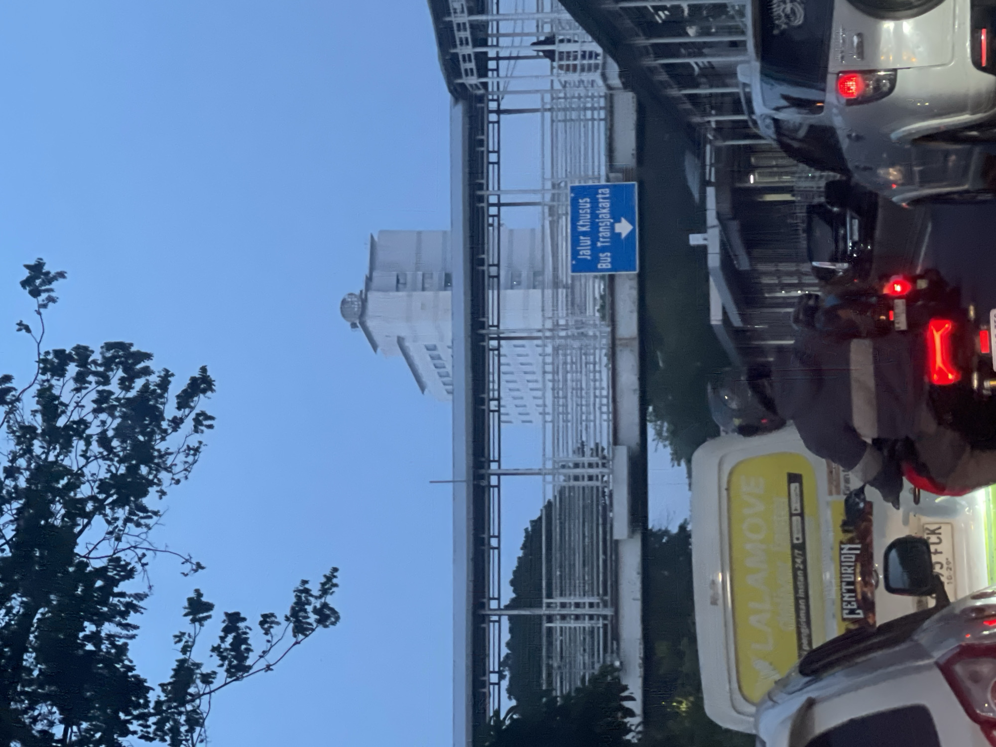 The image captures a street scene in Central Jakarta, Indonesia, likely during twilight or overcast conditions, given the muted lighting. A busy road is depicted with several vehicles, including cars, a van, and a motorcycle with a rider. Overhead, a pedestrian bridge spans across the road, with a blue sign indicating "Jalur Khusus Bus Transjakarta" (Special Lane for Transjakarta Bus) and a downward-pointing arrow. In the background, a tall, white building with multiple stories is visible, possibly a hotel or an office building. The foreground is partially obscured by the branches of a tree and the vehicles themselves. The overall impression is one of urban traffic, typical of a bustling city.