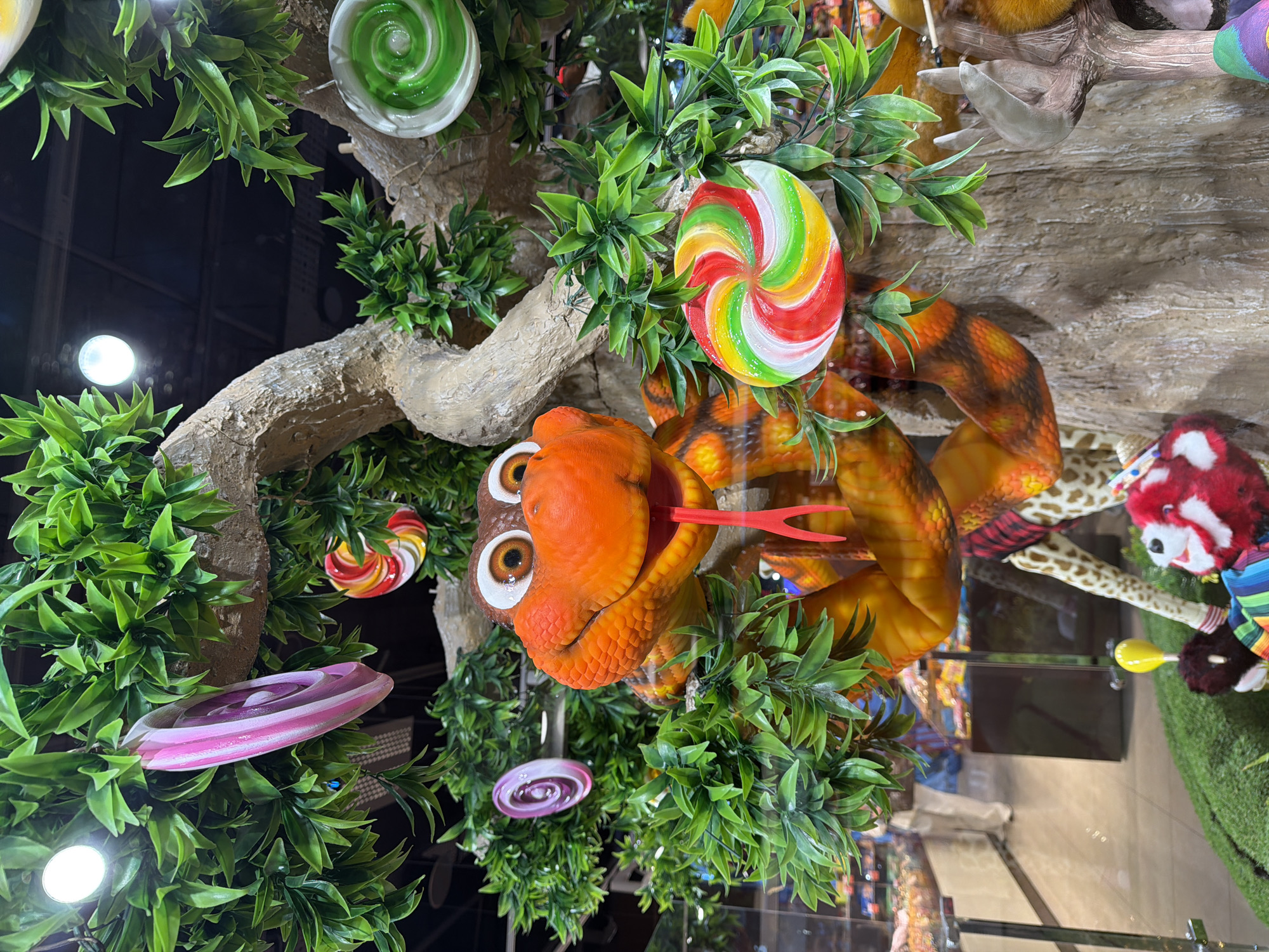 The image captures a whimsical display, likely at a confectionery or a themed attraction, in Sokilnyky, Ukraine. A prominent feature is a tree-like structure adorned with artificial green leaves. Hanging from the branches are several large, colorful, swirl-patterned lollipops, resembling those found in candy shops. These lollipops are pink and white, red, yellow, green, and white, and green.
A central focus is an animated, orange snake sculpture with large, wide eyes and a protruding red tongue. The snake's body is coiled around the tree trunk and branches, its scales detailed with patterns of orange, brown, and yellow.
In the background, through a glass pane, a store interior is visible. Shelves stocked with various candies are apparent, along with a person in casual attire walking in the distance, suggesting a retail environment. The foreground also shows portions of other plush toy-like decorations on a green, grassy base, hinting at a playful and festive setting. The lighting suggests an indoor environment, possibly a well-lit shop or exhibition space.