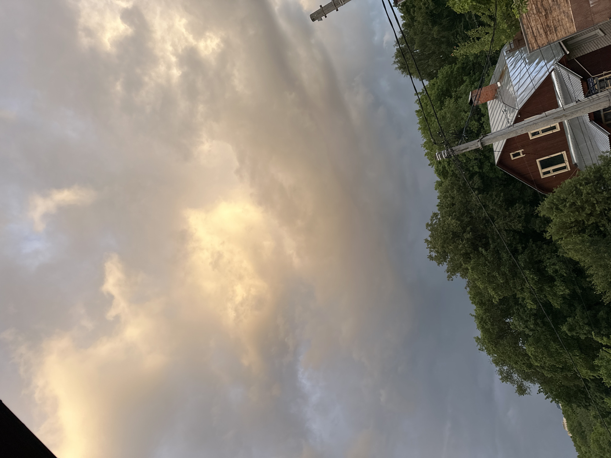 The image shows a view of the sky with heavy clouds, some lit by a warm golden light, likely from the setting sun. To the right of the frame, there's a building with a rusty-red facade and a metal roof, situated amidst green trees. An utility pole with electrical wires runs down the right of the building, slightly obscuring it. The background suggests a suburban or rural setting within the city limits of Nizhny Novgorod, Russia.