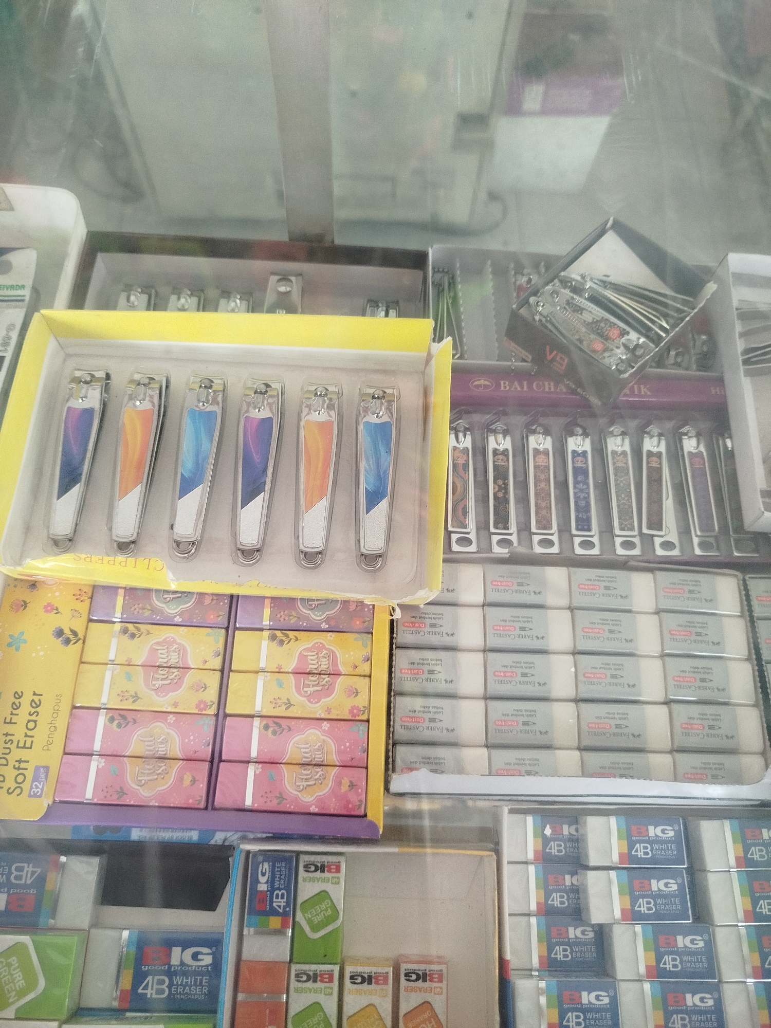 The image displays a retail display of stationery and personal grooming items, likely in a shop or market stall. The items include several rows of nail clippers, some with colorful handles and others with patterned designs. Below the nail clippers are stacks of erasers, some individually packaged and others in larger boxes. The erasers appear to be from brands like "Faber-Castell" and "BIG". There are also boxes of what look like scented erasers with floral designs. In the background, other items are visible, including what appears to be a set of multi-tools or knives in a black box, and possibly more stationery. The display is set within a glass-fronted cabinet or display case, suggesting an indoor retail environment. The lighting appears to be artificial, casting reflections on the glass. The specific location of Mranggen, Indonesia, cannot be determined from the visual information provided in the image. There are no people or interactions depicted.