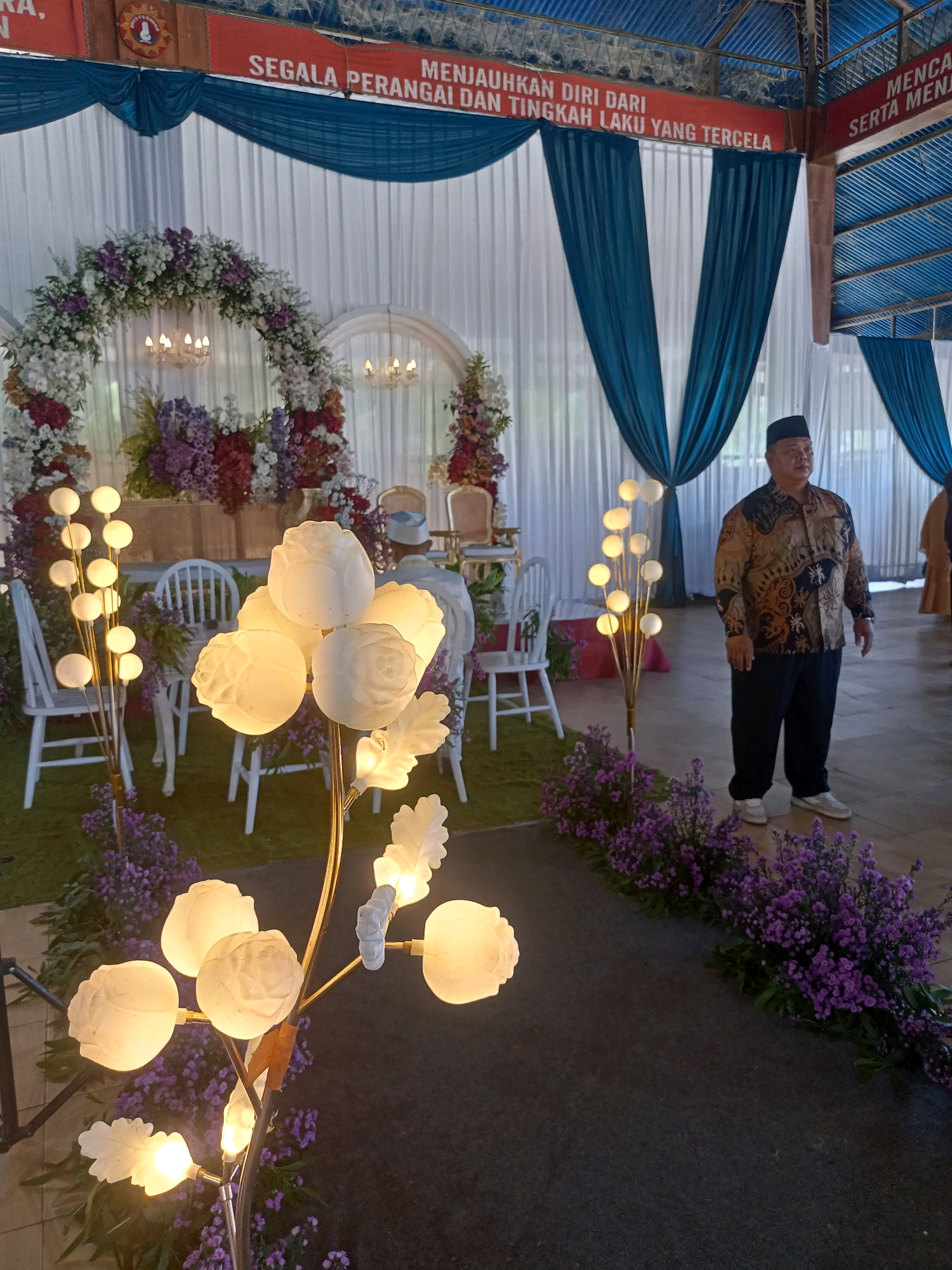 The image depicts an indoor event, possibly a wedding reception, set up in Wanadadi, Indonesia. In the foreground, a decorative lamp with multiple floral-shaped lights is prominently displayed. Behind it, a man in traditional Indonesian attire, a batik shirt and dark pants with a peci hat, stands facing left. He appears to be observing the surroundings. The background features a stage area decorated with a large floral arch composed of white and purple flowers, white drapes, and blue curtains. Several white chairs are arranged around the stage. Above the stage, a red banner with Indonesian text is visible. The flooring appears to be made of dark carpet or asphalt. The overall atmosphere suggests a formal gathering or ceremony.