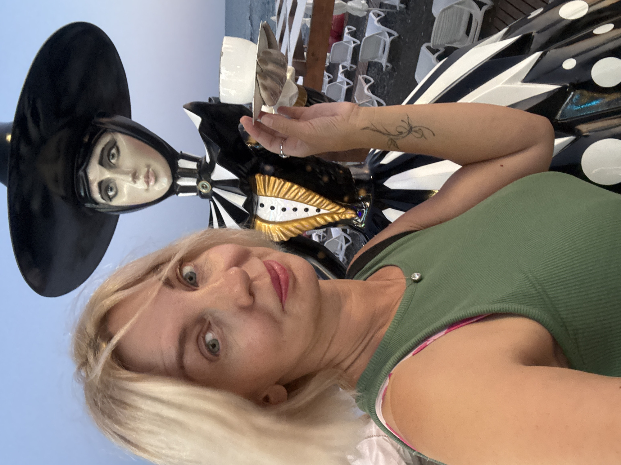 A selfie taken by a woman with blonde hair and a green tank top at a beach resort in Sochi, Russia. She is standing next to a life-sized statue of a person dressed in a black and white outfit, with a large black hat and a golden waistcoat. The statue is holding a tray with a white cup and saucer. In the background, there are rows of white lounge chairs on a pebble beach, with the Black Sea and the sky visible. The overall lighting suggests it is evening.