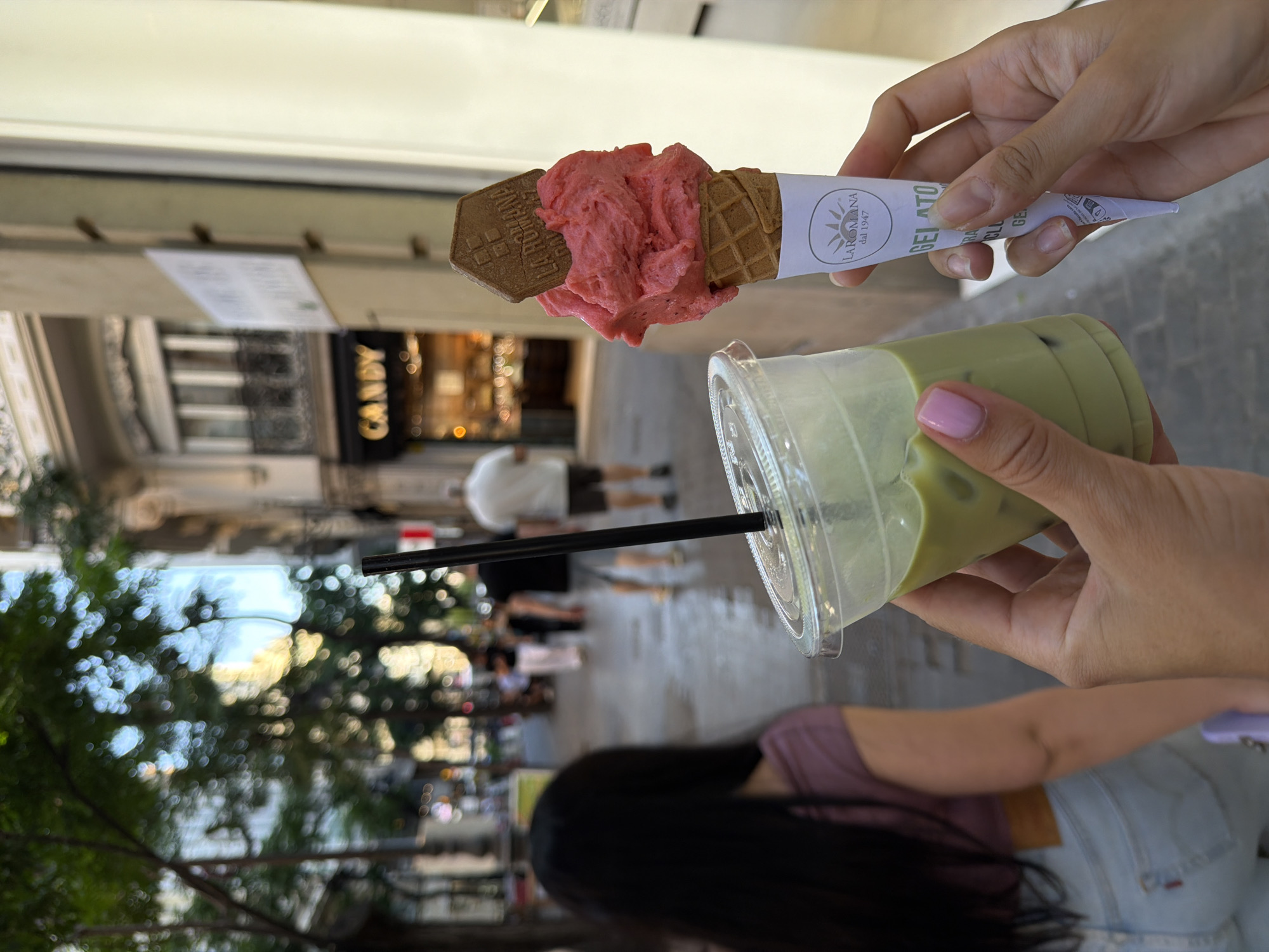 In this medium shot, a person's hands hold a strawberry gelato cone and a clear plastic cup of iced matcha latte on a city street. The gelato is a vibrant pink and topped with a rectangular wafer with a logo. The cup contains a green beverage with ice and a black straw. In the background, a woman with long dark hair and a purple shirt walks away from the camera, wearing light-wash jeans. Further down the street, blurred figures of people are visible, some walking, and a store sign that reads "CANDY." The environment appears to be a commercial area with trees and buildings lining the street.