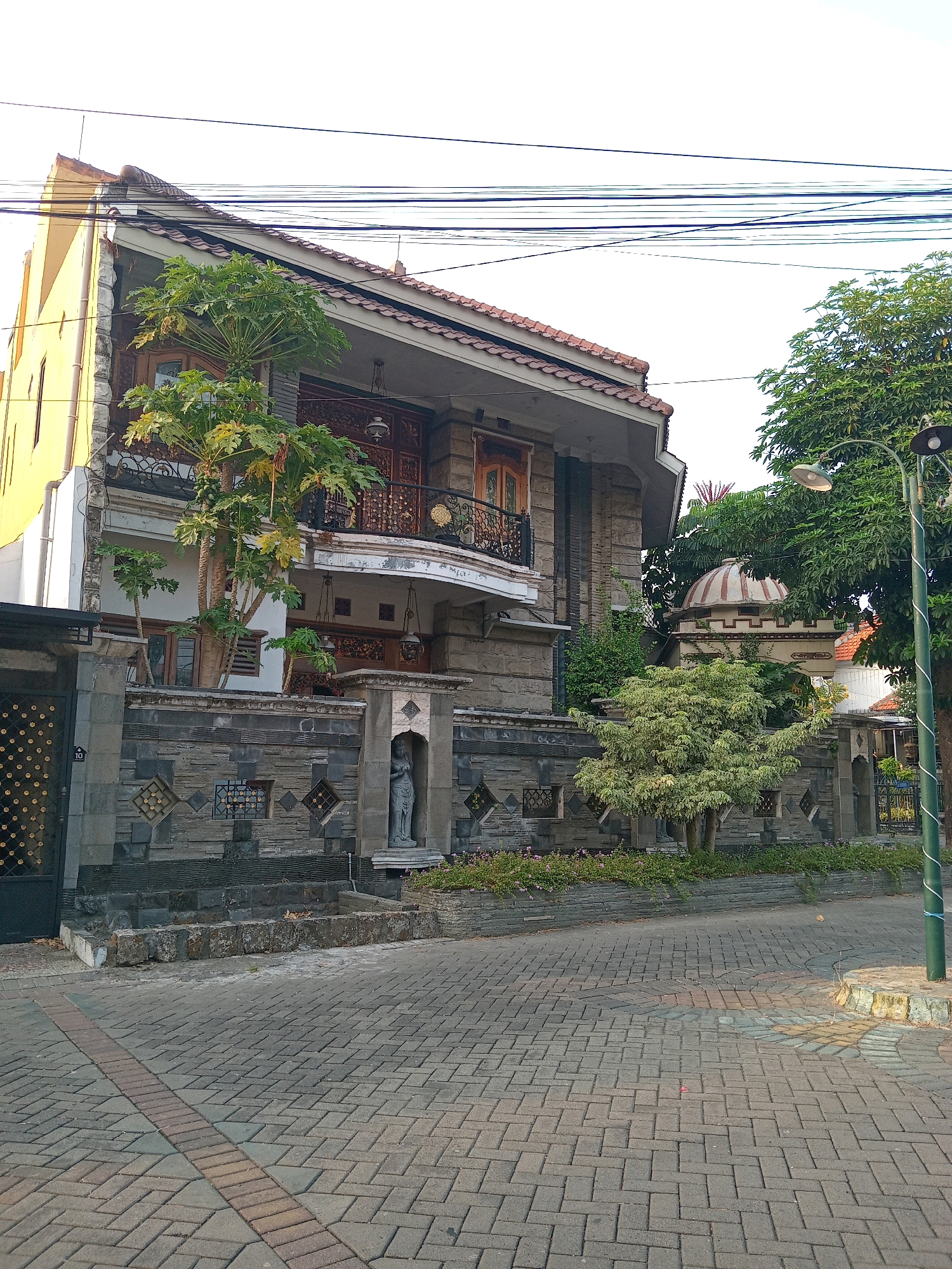 In Randuagung, Indonesia, a two-story house with a yellow facade and stone accents stands at the end of a paved driveway. The house features a prominent balcony with ornate railings and a tiled roof. A papaya tree grows beside the house, its leaves reaching up towards the balcony. A stone wall with decorative geometric patterns encloses the property. Within an alcove in the wall stands a grey statue of a woman. A small domed structure, possibly a gazebo or folly, is visible behind the wall. Green trees and a streetlamp are also present. The sky is overcast.