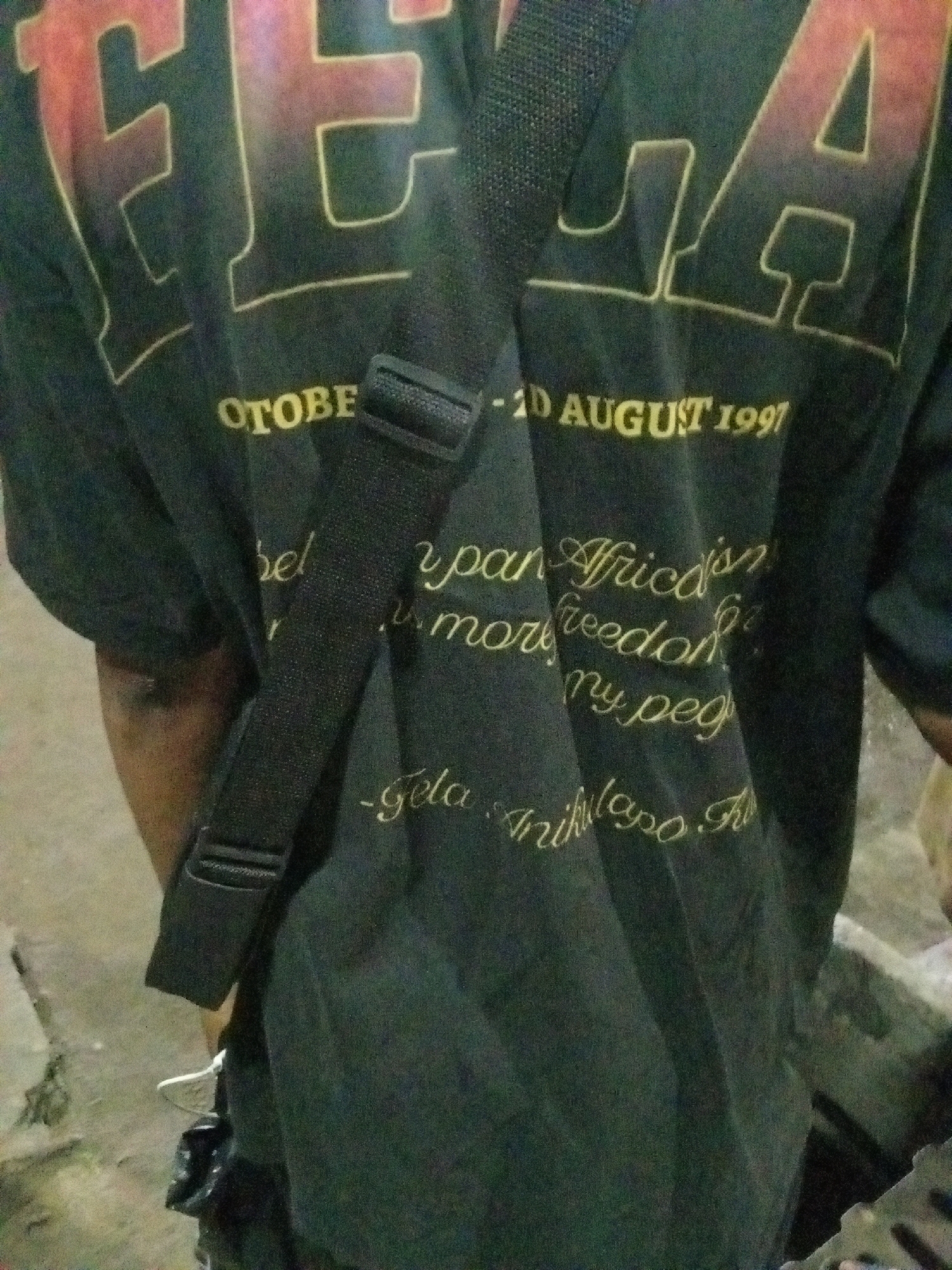 A man stands with his back to the viewer, wearing a dark t-shirt. The t-shirt has a design with the words "FELA" in large, outlined letters at the top, followed by smaller text: "OCTOBER...-2D AUGUST 1997". Below this, more text is visible, including "Pan Africa", "freedom", and "Anikulapo". A black strap crosses over his shoulder and back. The environment appears to be an urban setting in Ado, Nigeria, given the provided information.