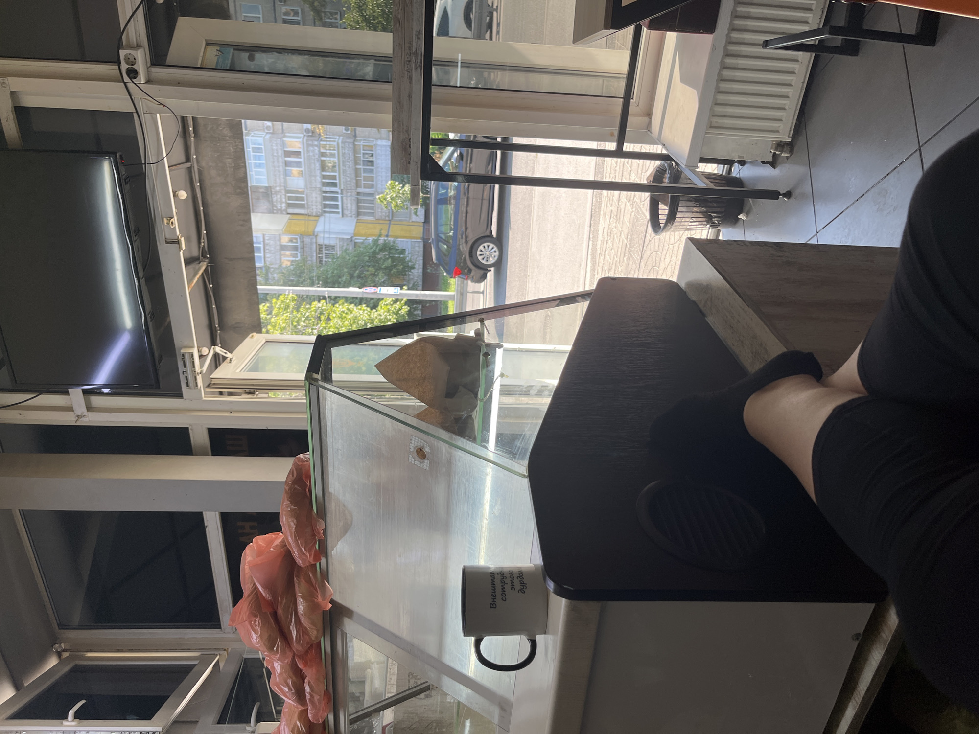 The image shows a person's legs crossed, resting on a dark brown wooden counter in what appears to be a small shop or cafe, possibly in Dnipro, Ukraine. A white mug with black lettering sits on the counter to the left. In the background, a glass display case filled with food items is visible. Beyond the shop, an urban street scene is visible with buildings and a black car driving on the road. The lighting suggests it is daytime.