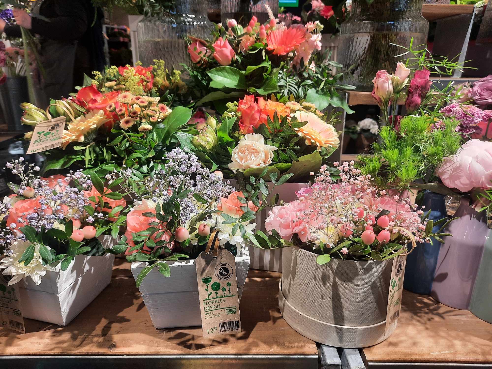 The image depicts a display of floral arrangements on a wooden surface, likely within a retail setting. Several bouquets are visible, featuring a variety of flowers in shades of pink, orange, coral, and cream, interspersed with greenery. Some arrangements are presented in white wooden boxes, while others are in a textured, light-colored cylindrical container. A prominent tag on one of the white boxes displays "Florales Design" and "Mala 2" along with a barcode and price. In the background, glass vases and blurred figures of people can be discerned, suggesting a flower shop or market. The lighting appears to be artificial, illuminating the colorful blooms. The overall scene conveys a commercial presentation of fresh flowers. The image is not geotagged and therefore the exact location in Rosenheim, Germany cannot be determined.