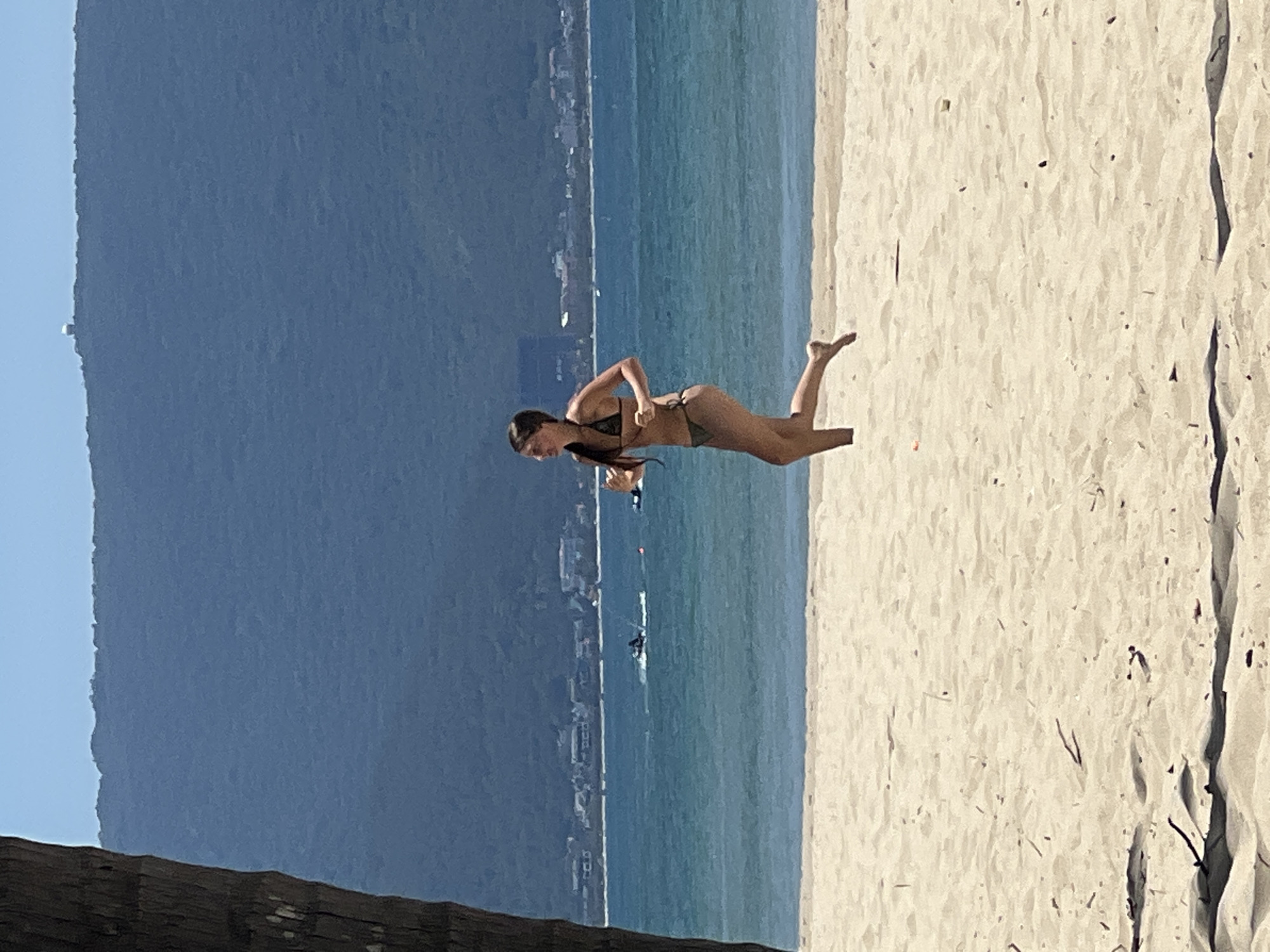 A woman in a dark green bikini is running along a sandy beach. She is facing to her left and her hair is tied back in a ponytail. Behind her, the turquoise ocean stretches out, with a faint line of buildings and a mountainous backdrop. A palm tree trunk is visible on the far left of the frame. The scene appears to be set in Da Nang, Vietnam, possibly along the coastline with views of the Son Tra Peninsula.