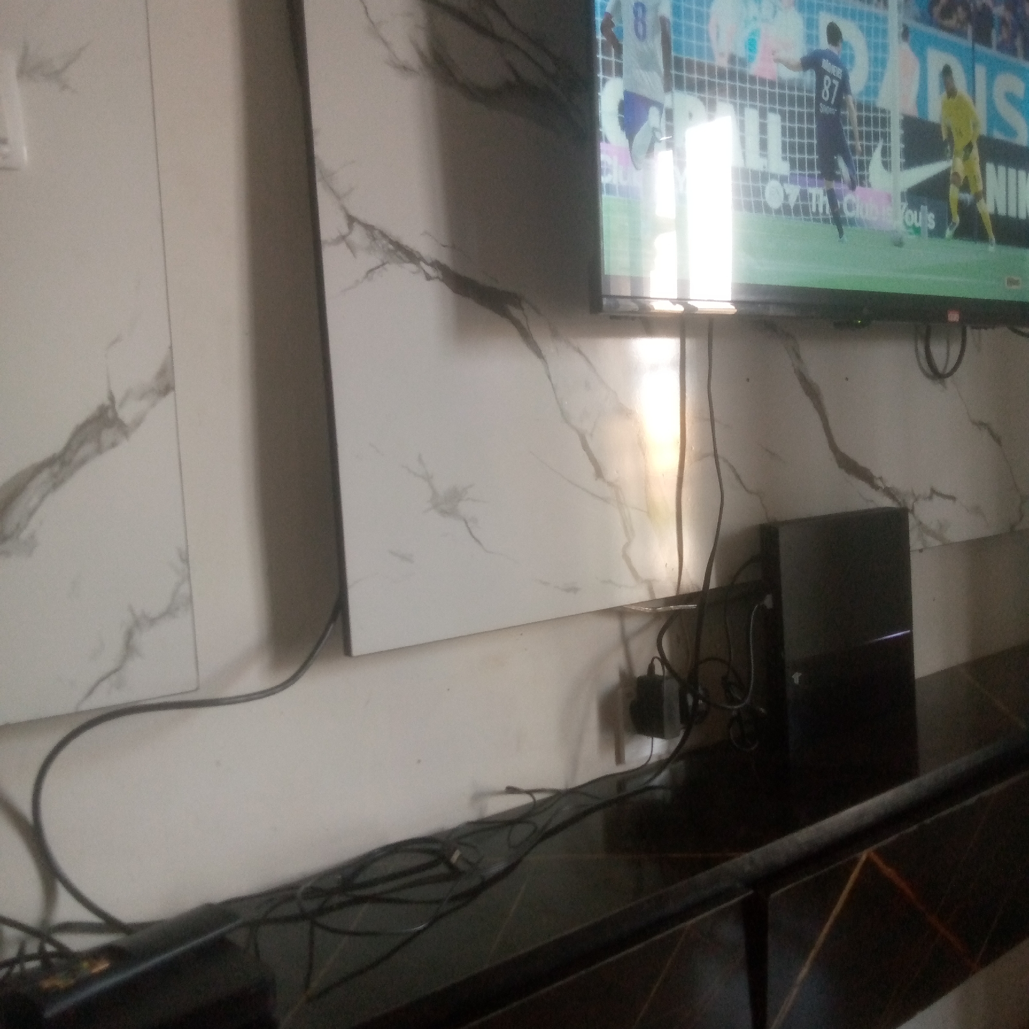 The image depicts a television screen displaying a soccer game, with players in blue and yellow uniforms on a green field. The television is positioned above a dark furniture piece. To the left of the television, a portion of a wall with a marble-like pattern is visible. On the furniture piece below the television, a black electronic device, likely a game console, is present along with several black cables. One cable is plugged into a black power adapter mounted on the wall behind the console. The environment appears to be indoors, with the lighting suggesting it could be a living room or entertainment area. The location is identified as Unknown, Unknown.