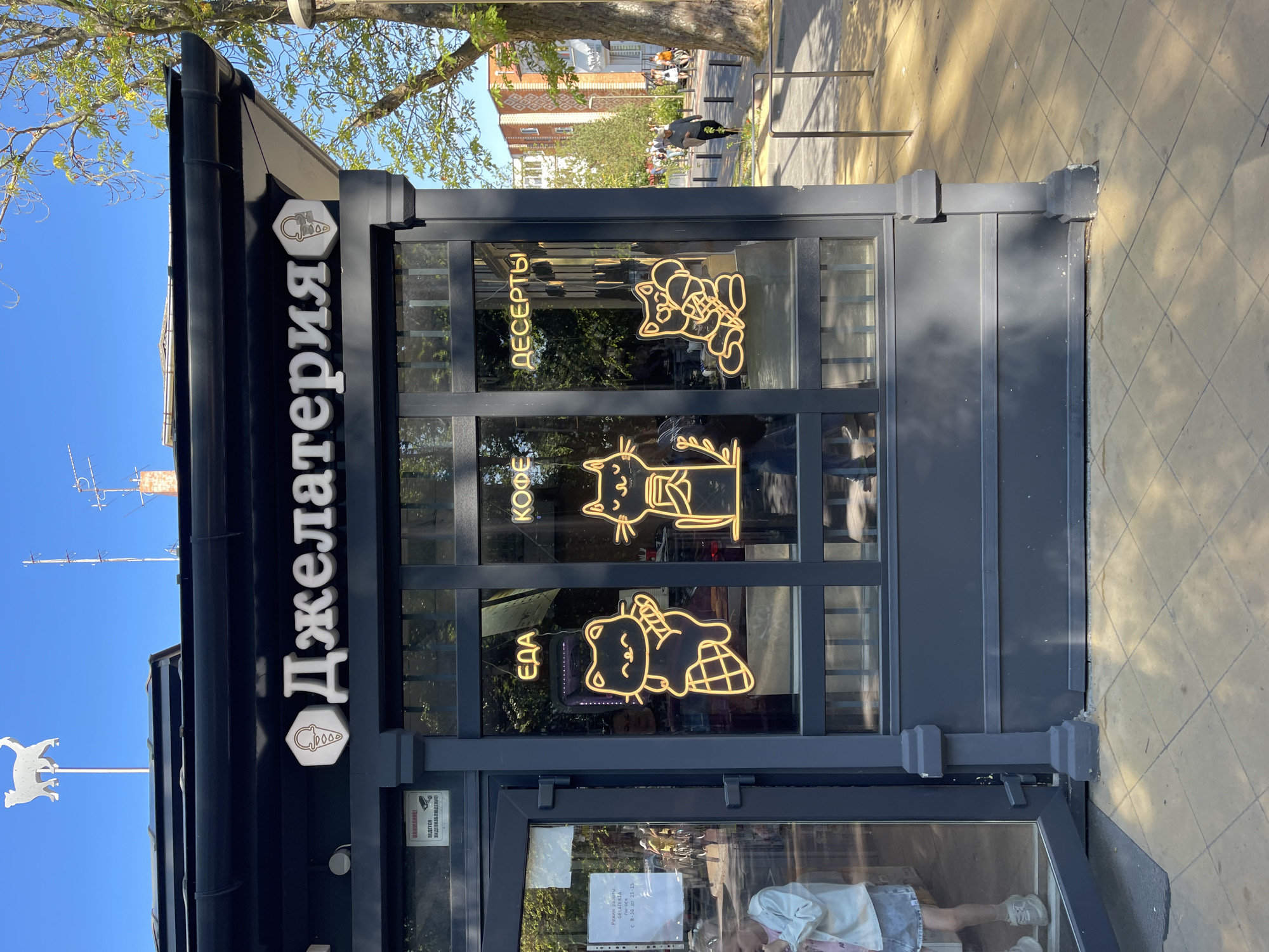 This media file shows the exterior of a gelato shop called "GELATERIA" in Zelenogradsk, Russia. The shop is a small, dark gray building with large glass windows. Through the windows, neon signs of cartoon cats can be seen, advertising "ЕДА" (food), "КОФЕ" (coffee), and "ДЕСЕРТЫ" (desserts). Above the window, the shop's name, "Джелатерия," is displayed in large white letters, with a small ice cream cone logo. A white metal weather vane shaped like a cow is visible on a pole to the left of the building. The image is taken during the daytime under a clear blue sky, with sunlight casting shadows on the pavement in front of the shop. The pavement is paved with square tiles. In the background, there are trees and buildings, and a few people are walking on the sidewalk. One sign inside the shop indicates its operating hours from Monday to Sunday, 8:30 AM to 9:15 PM. Another sign warns that video surveillance is in progress. The overall scene suggests a bright and sunny day in a pleasant urban environment.