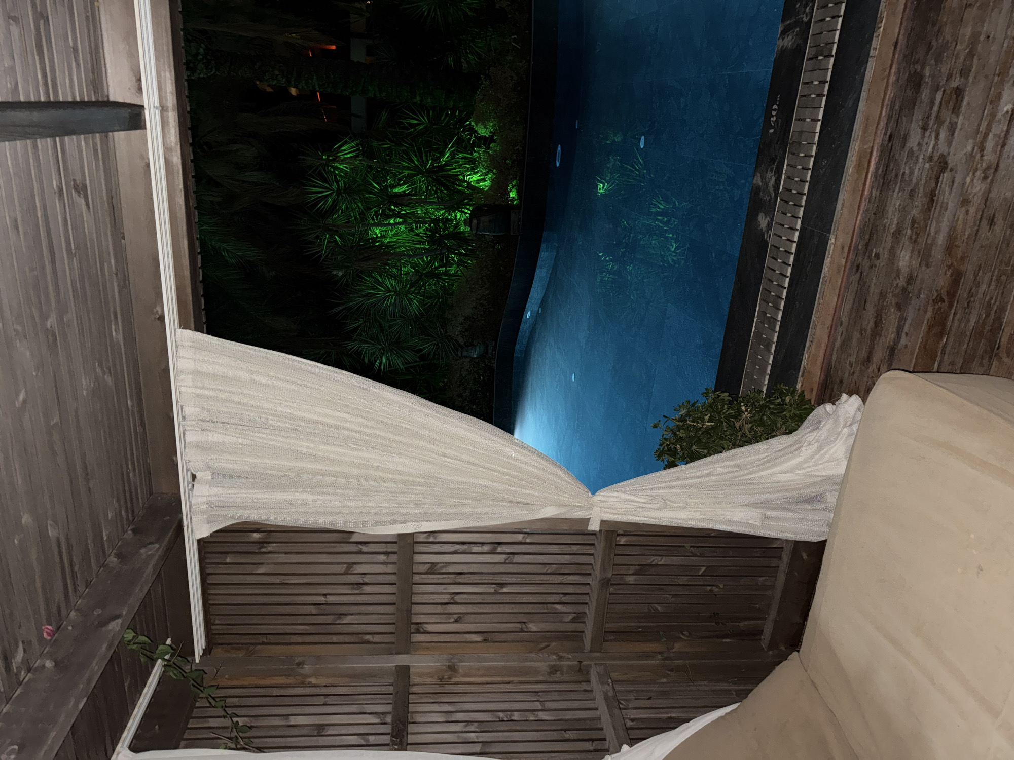 The image depicts a night scene at a resort in Bodrum, Turkey. The viewpoint is from a covered outdoor lounge area, looking out towards a swimming pool. The lounge area has a wooden slatted ceiling and wooden paneled walls. A light-colored, sheer curtain is drawn partially across the opening, tied back with a knot. To the left, a portion of a tan-colored daybed is visible.
Beyond the lounge, a bright blue swimming pool is illuminated. The water reflects the light, and subtle circular disturbances are visible on its surface. The pool is bordered by a darker material with a slatted drain. In the background, lush green foliage is heavily illuminated by bright green lights, creating a vibrant, almost artificial glow against the dark night sky. Palm trees and other dense vegetation are discernible within this illuminated area. The overall impression is one of a tranquil, possibly luxurious, nighttime setting.