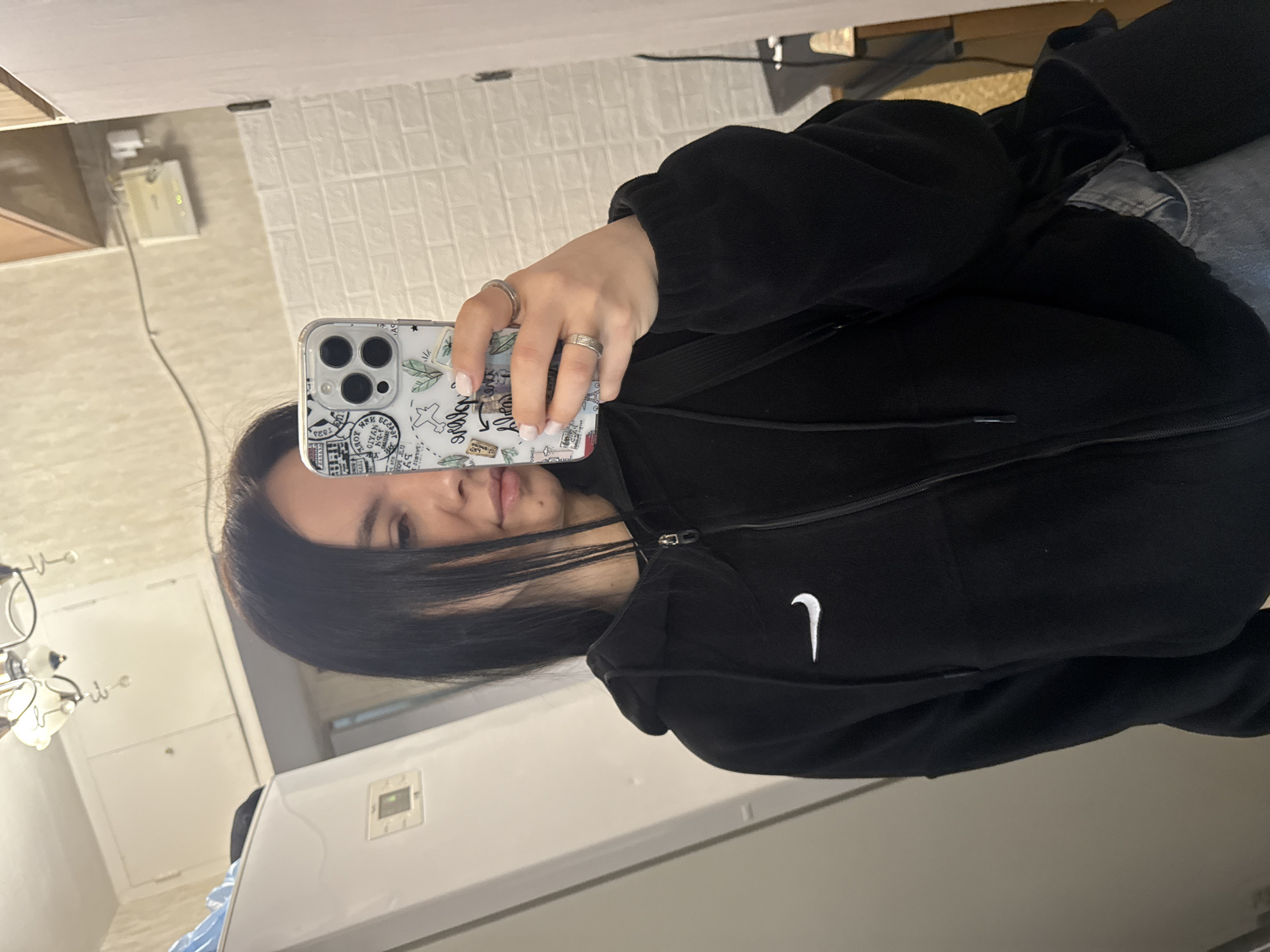 A person with long dark hair is holding a smartphone with a patterned case and silver rings on their fingers, taking a selfie in front of a mirror. The person is wearing a black Nike hoodie and blue jeans. The background shows a portion of a room with white walls and a glimpse of kitchen cabinets. The reflection in the mirror reveals a white cabinet and a light fixture. The location in Mytishchi, Russia is not apparent from the image.