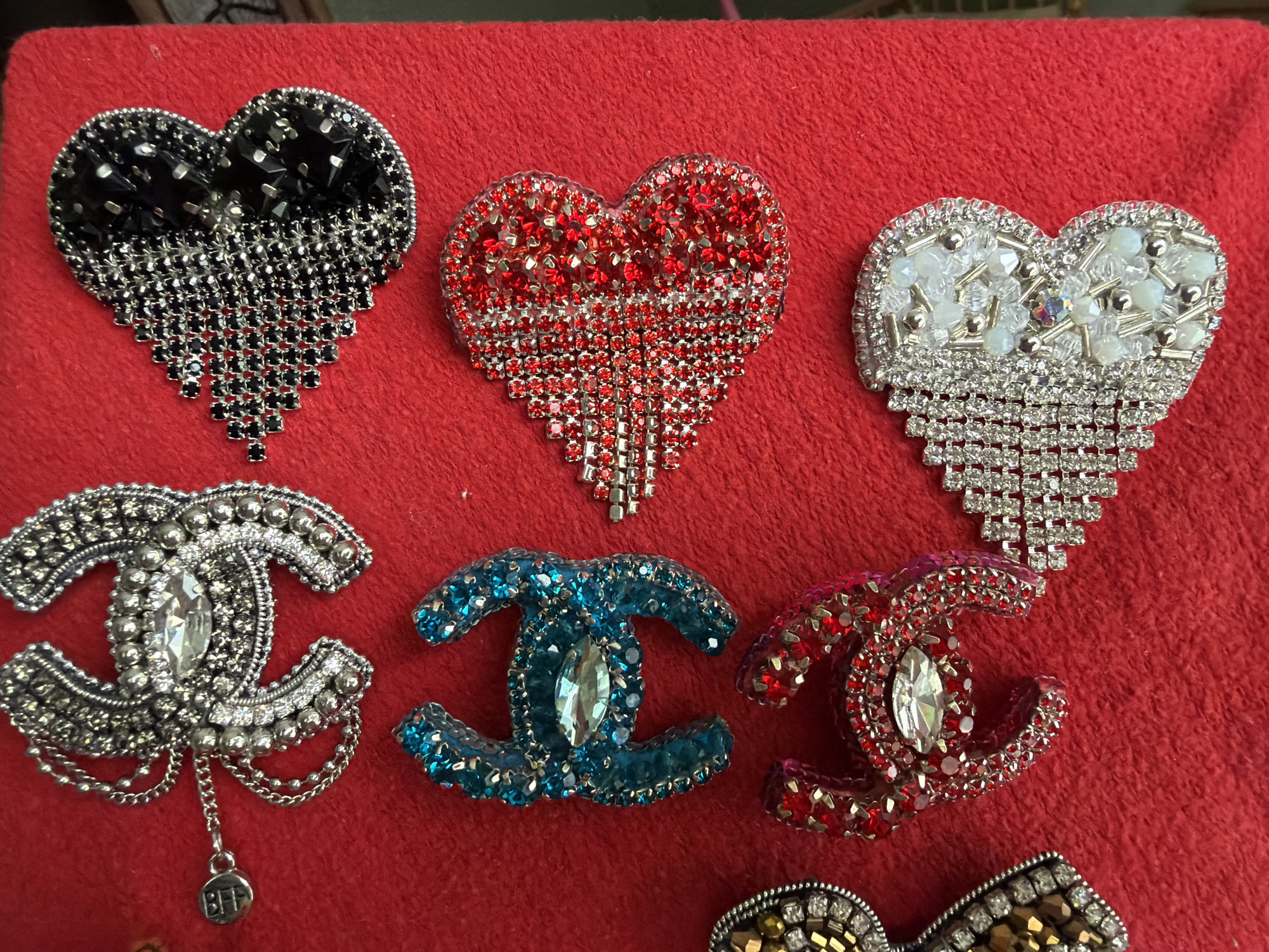The image shows several decorative brooches and emblems arranged on a red fabric surface. The items include three heart-shaped brooches, each adorned with different colored gemstones or beads (black, red, and white). Below these, there are three emblems resembling the interlocking "C" logo, embellished with silver, blue, and red gemstones, respectively. A silver brooch with a dangling charm is also present. The environment suggests a craft or jewelry display setting.