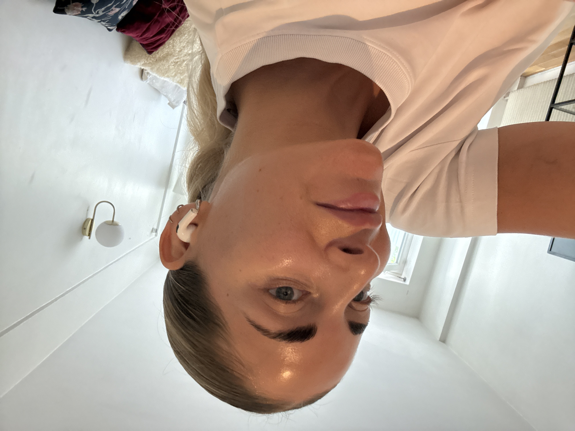 The image features a close-up, inverted shot of a young woman's face and upper body, positioned against a clean, white wall. She has blonde hair pulled back, showcasing her face with fair skin that appears to have a dewy or glowing finish. Her eyes are a striking blue-grey, and she has well-defined eyebrows. She is wearing a white t-shirt. In her right ear, she has a white wireless earbud.
The background is simple and suggests an indoor setting. To the left, a small, modern wall-mounted light fixture with a white globe shade is visible. Further in the background, a window is partially visible, and to the right, a black metal structure, possibly part of furniture or a shelf, can be seen. The lighting suggests it could be daytime, with natural light possibly coming from the window. The overall mood is casual and relaxed. The woman's gaze is directed slightly away from the camera, conveying a neutral or thoughtful expression. No text is visible in the image.