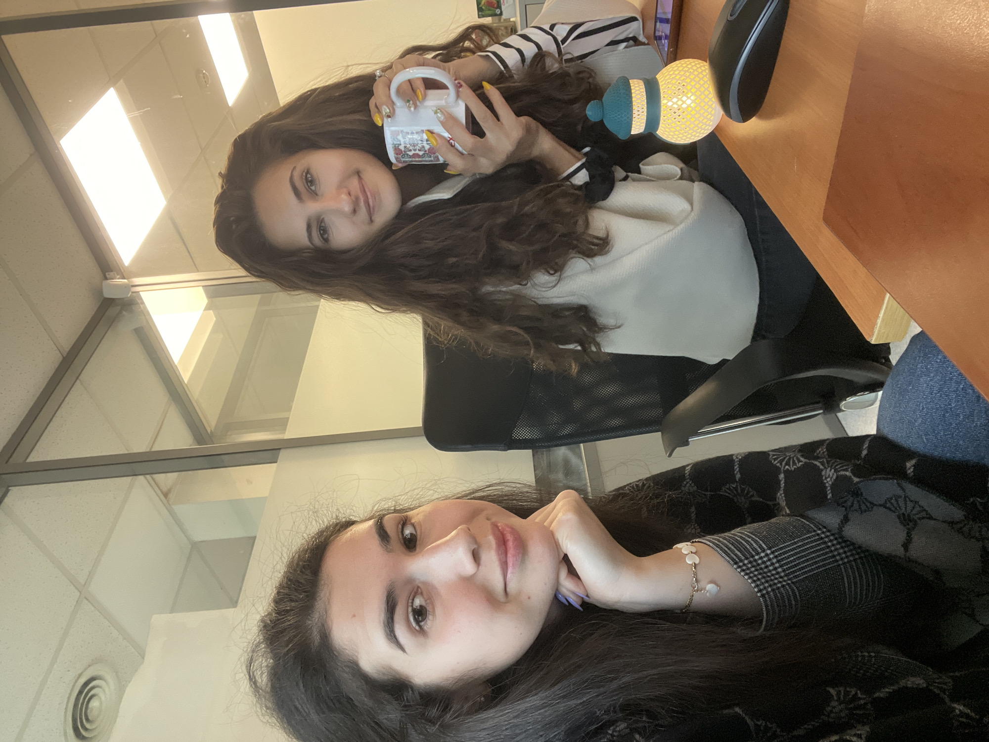 Two women pose in an office setting. The woman in the upper portion of the frame is seated at a desk and holding a mug with decorated nails. A lamp and computer mouse sit on the desk beside her. She is wearing a striped shirt and white sweater. The woman in the lower portion of the frame is resting her chin on her hand. She has a bracelet on her wrist and is wearing a patterned shirt. The location is likely an office building in Sofia, Bulgaria, given the architectural style of the ceilings and windows.