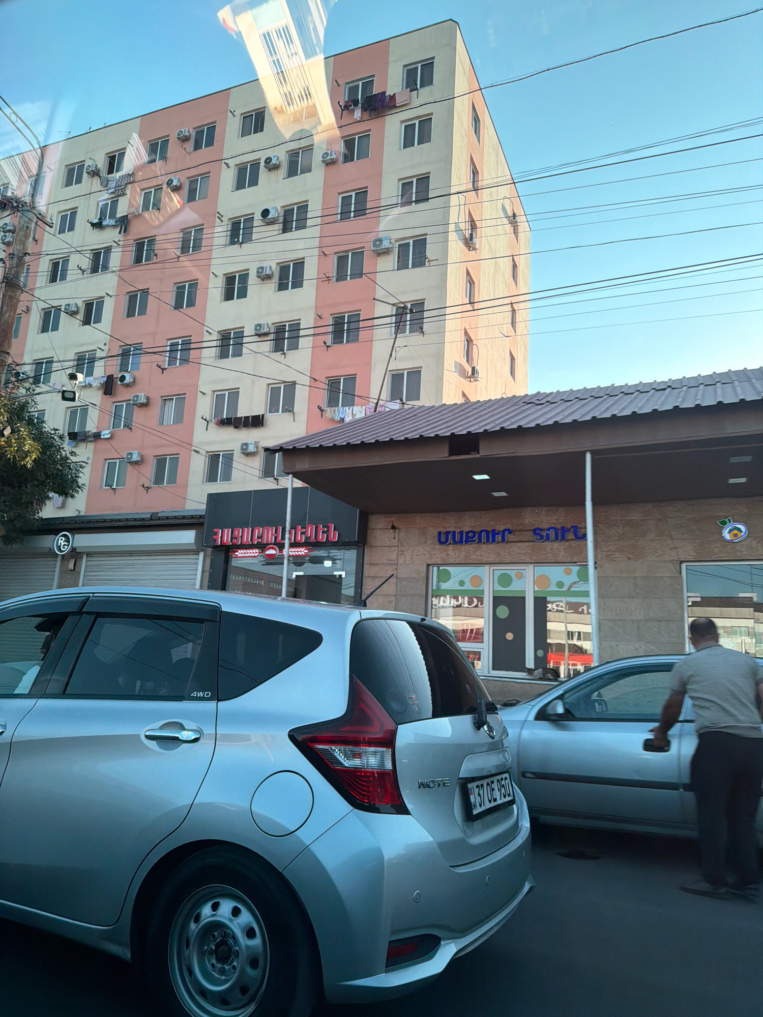 A silver Nissan Note is parked on the street in Yerevan, Armenia. The car is facing towards the right of the frame. In the background, a light-colored building with peach-colored accents rises several stories high and is covered in numerous air conditioning units. A lower building with a dark roof and beige facade is in front of the taller building, featuring storefronts. One storefront has blue text that reads "ՄաՔարո Սաֆթ" (Makar Soft). Another storefront has red text that reads "Հայաստան Բանկ" (Hayastan Bank). Clothes are hanging from lines on the exterior of the taller building. A man wearing a grey t-shirt and dark pants is standing next to a silver sedan, reaching towards its door handle. The sky is clear and blue. The scene appears to be a street in a residential or commercial area of Yerevan.