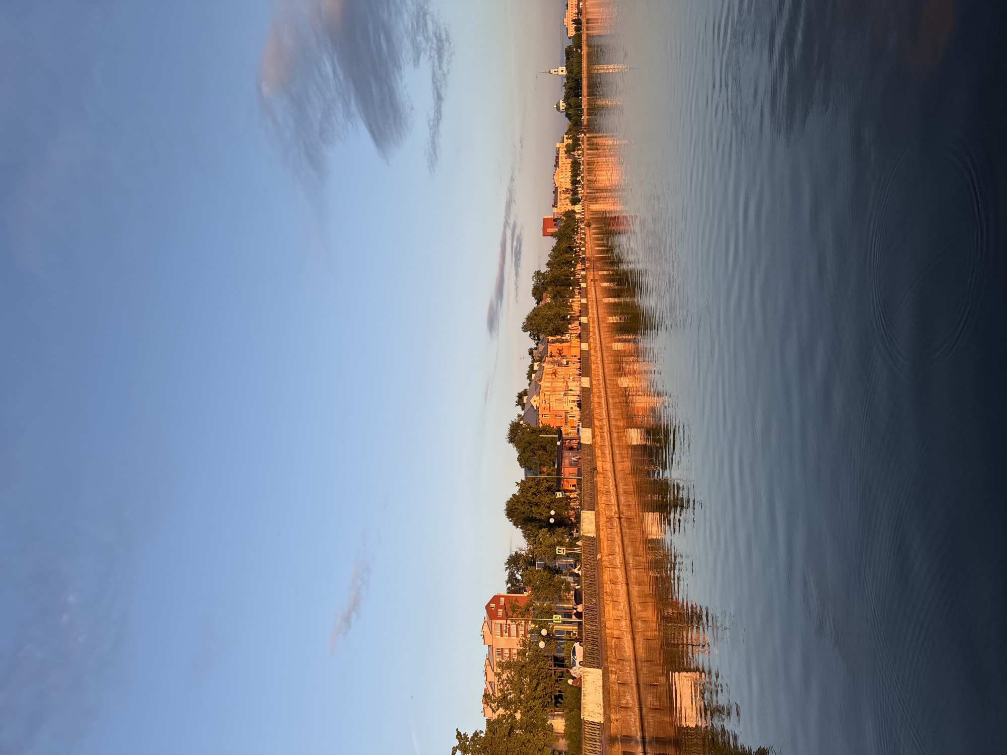 This image, when viewed with the correct orientation (rotated 90 degrees counter-clockwise), depicts a serene panoramic view of a city embankment alongside a body of water, likely in Votkinsk, Russia. The scene is illuminated by the soft, warm light of late afternoon or early evening, creating a tranquil atmosphere.
The upper portion of the frame shows a clear, pale blue sky adorned with a few wispy, elongated clouds, some touched by the warm, golden hues of the setting or rising sun. Below the sky, the city's embankment stretches horizontally across the middle of the image. A variety of buildings, featuring different architectural styles from classical to more contemporary, line the waterfront. Many structures appear to be bathed in warm, golden light, casting rich ochre and reddish tones. Green trees with dense foliage are interspersed along the embankment, partially obscuring the buildings. Streetlights and some unreadable yellow road signs are visible along the path that runs parallel to the water. In the far distance, a small boat or its mast can be faintly seen.
The lower half of the image is dominated by the calm water, which acts as a mirror, reflecting the sky, the buildings, and the golden light. These reflections are beautifully distorted by gentle ripples and small waves on the water's surface, creating an abstract, shimmering effect. On the right side of the water, distinct concentric ripples are visible, indicating a recent disturbance to the otherwise calm surface. The water transitions from lighter, reflective areas near the shore to darker, deeper blues further out. There are no discernible people or specific activities taking place, emphasizing the peaceful and still nature of the scene.