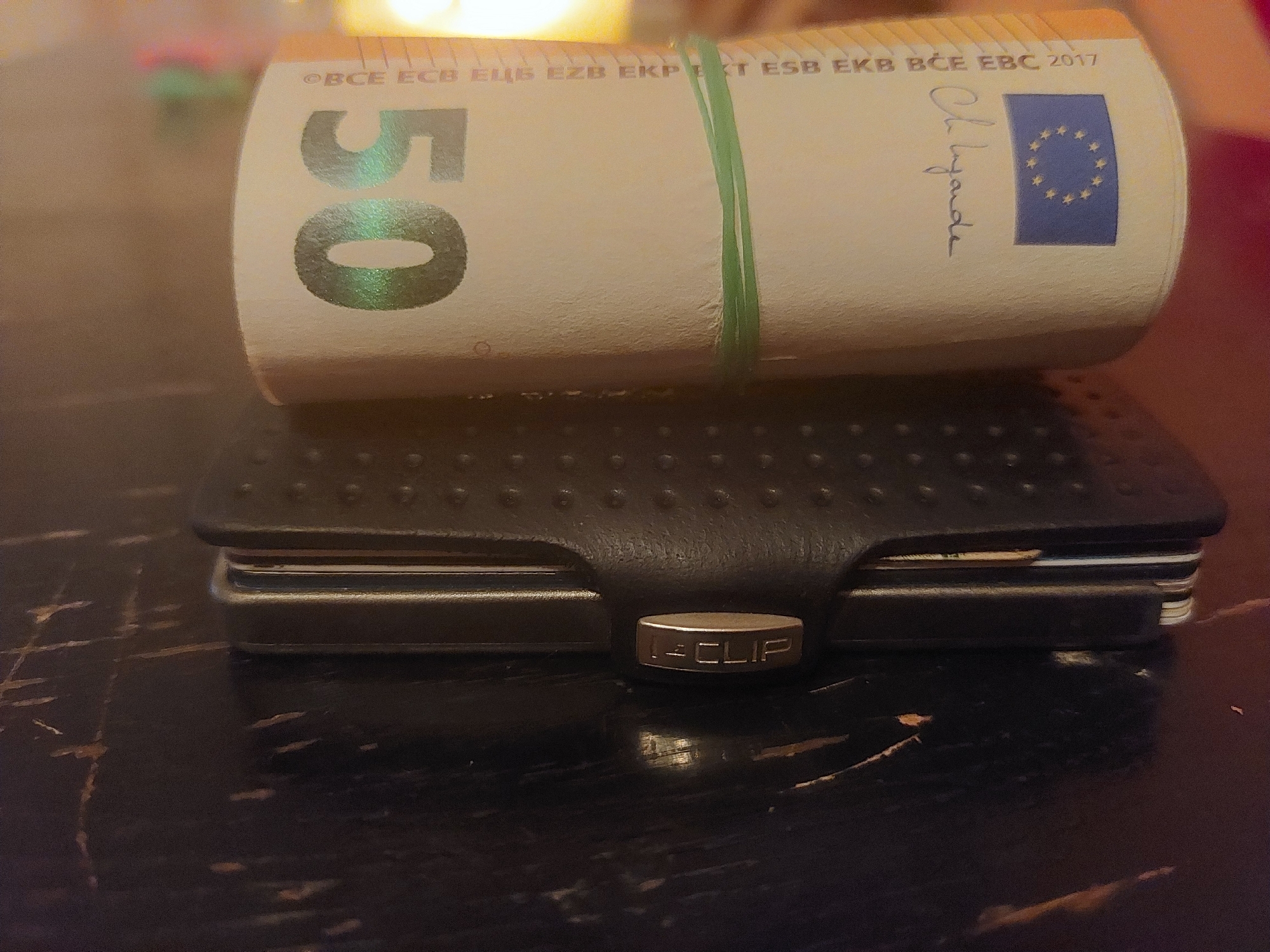 The image shows a roll of 50 Euro bills secured with a green rubber band, sitting on top of a black I-CLIP wallet. The bill has the number "50" visible on one end, and the Eurozone flag and "2017" printed on the other. The I-CLIP wallet appears to hold several cards. The objects are placed on a dark, scratched, reflective surface. The location in Tsarevo, Bulgaria is not evident in the image.