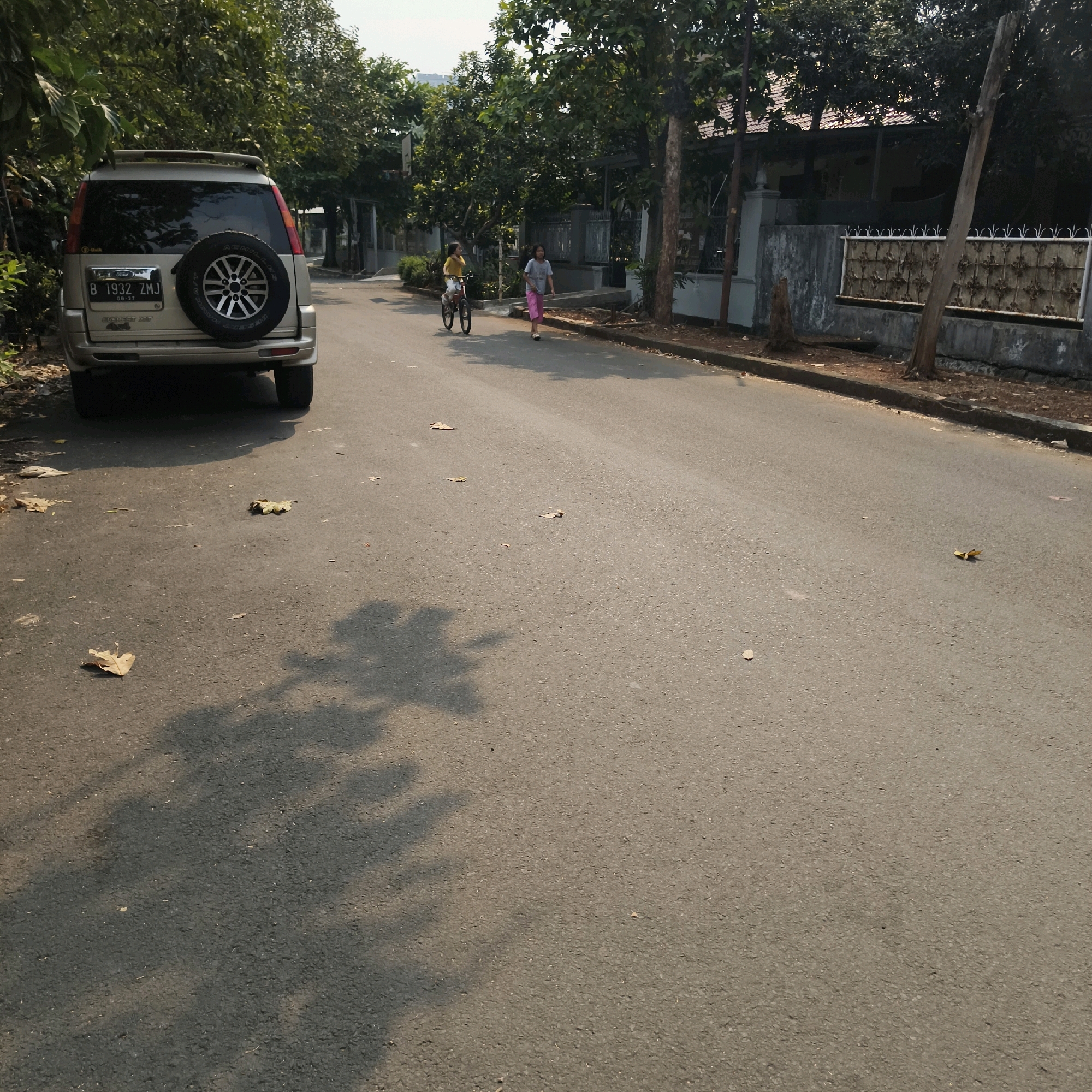 In South Jakarta, Indonesia, a street scene unfolds under daylight. A silver Ford Everest SUV is parked on the left side of the asphalt road. Further down the road, a young girl is riding a bicycle, wearing a yellow shirt. Beside her, another girl in a gray shirt and pink pants walks along the sidewalk. The street is lined with trees and residential houses with fences. Scattered dry leaves are visible on the road and the side of the street. Shadows from trees and possibly the SUV are cast across the asphalt.