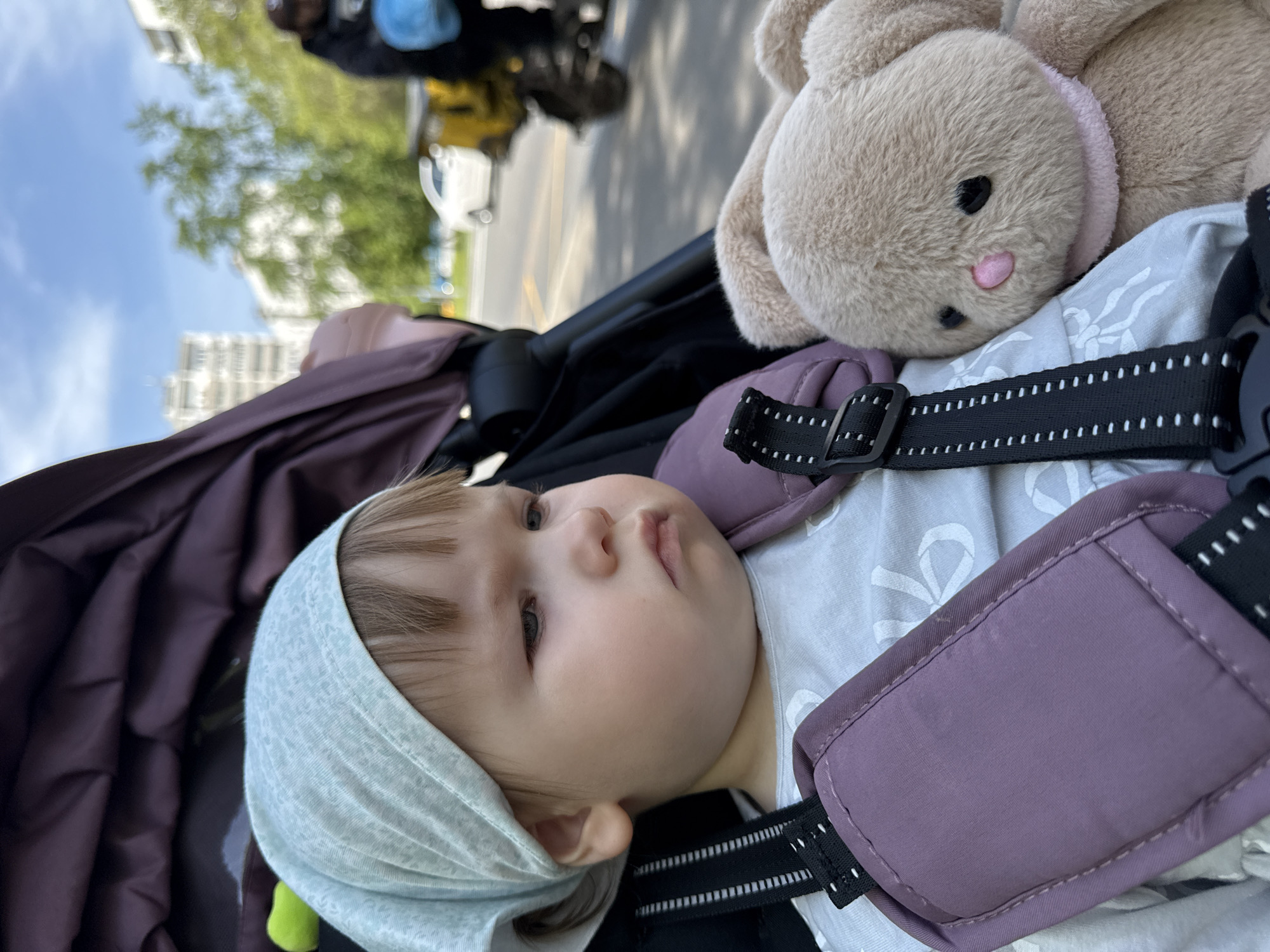 A child in a stroller is lying next to a stuffed bunny. The child is wearing a head covering, and the stroller has a safety harness. The background shows a street with a building and trees. The location appears to be a city street, potentially in Moscow, Russia. The stroller may be on the move.