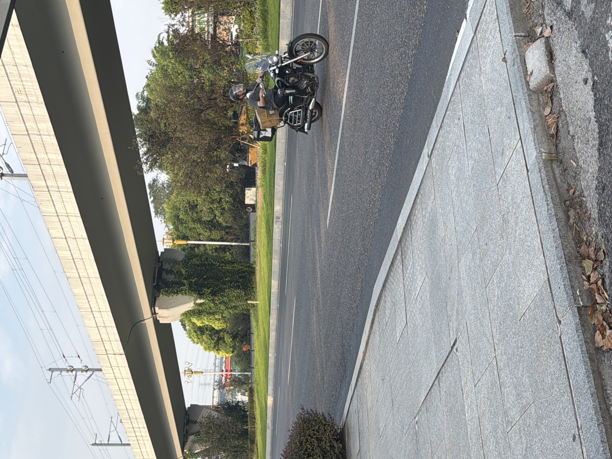 A man wearing a helmet and a light grey t-shirt is riding a black motorcycle on an asphalt road. He is carrying a brown cardboard box on the back of the motorcycle. The road is bordered by a grey concrete sidewalk with square paving stones and a small patch of brown leaves. In the background, there is a raised concrete structure, possibly a bridge or overpass, with electrical wires running above it. Green trees and foliage are visible behind the road. The sky is clear and blue. The scene appears to be an urban or suburban environment in Jiangjin, China.