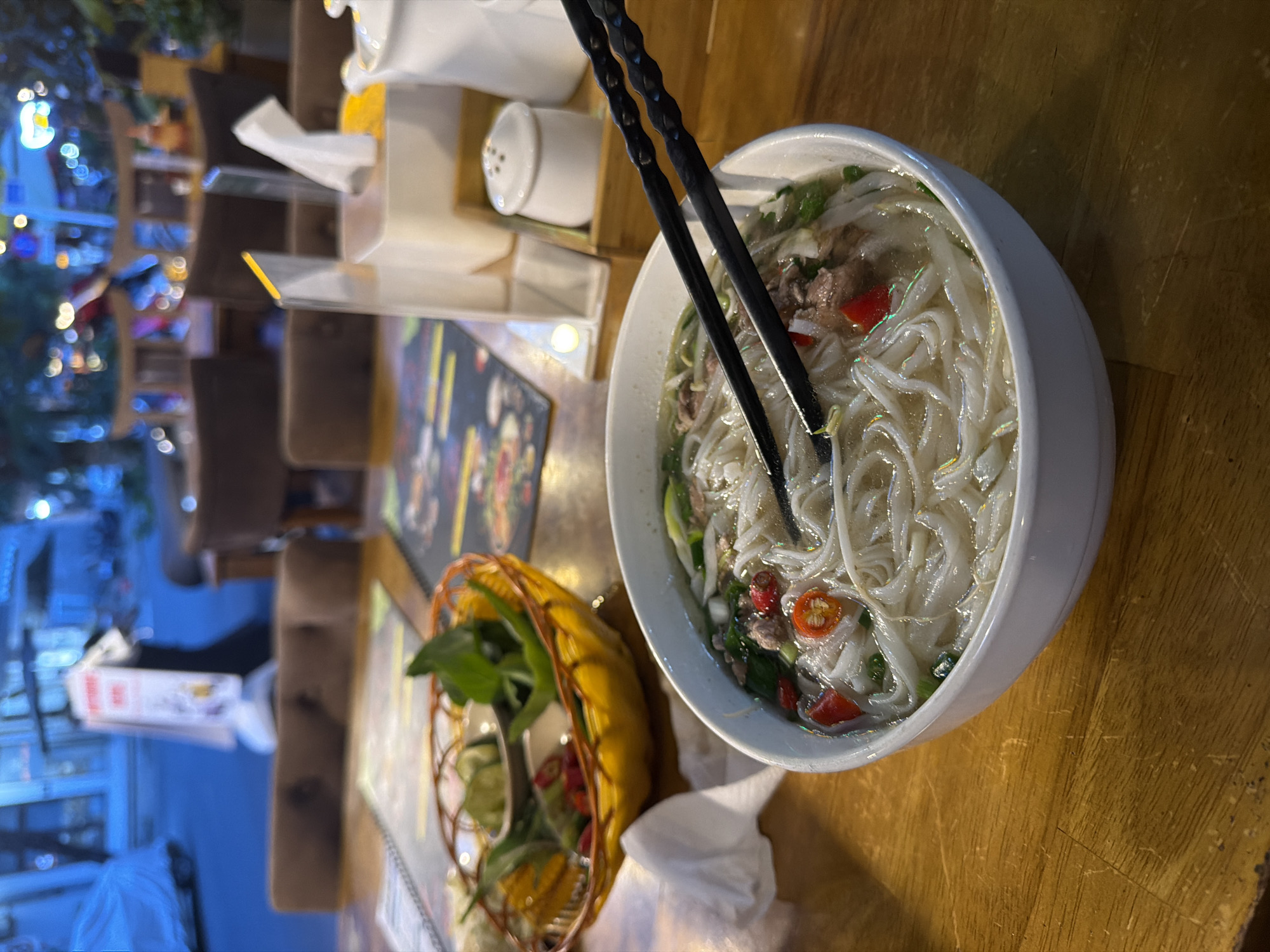 The image shows a bowl of pho, a Vietnamese noodle soup, on a wooden table. The bowl is white and contains rice noodles, broth, meat, green onions, and chili peppers. A pair of black chopsticks rest on top of the bowl. A small basket containing fresh herbs and lime wedges sits next to the bowl. In the background, there is a condiment set including a white box and salt shaker, with a restaurant menu visible partially. The blurred background suggests a restaurant setting in the city of Đà Nẵng, Vietnam, with city lights visible outside. No people are visible in the image.