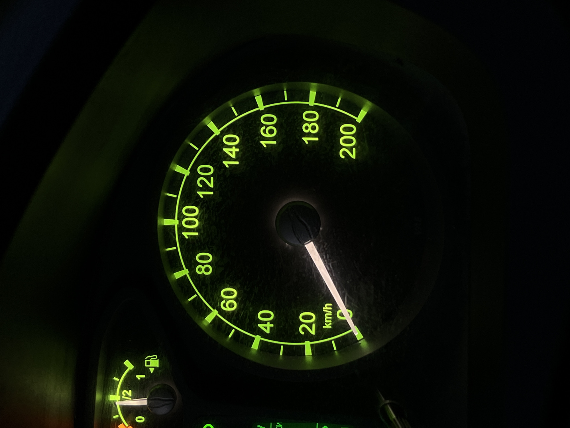 This is a close-up, dimly lit photograph of a car's dashboard, primarily featuring the speedometer and a fuel gauge. The dominant subject is the speedometer, a circular gauge with bright green markings and numbers illuminated against a dark background. The needle of the speedometer is pointing slightly past the 20 km/h mark, indicating the vehicle is moving at a slow speed. The numbers on the speedometer range from 0 to 200 km/h, with tick marks at 20 km/h intervals.
Below and to the left of the speedometer is a smaller, curved fuel gauge. Its needle is positioned between the "0" and "1/2" markings, suggesting the fuel tank is less than half full. The fuel symbol, a gas pump icon, is also visible near the gauge.
The overall scene is dark, suggesting it is either nighttime or the interior of a vehicle with its lights on. The green glow from the dashboard instruments is the primary light source in the image. The surface of the dashboard appears slightly dusty or textured. There are no people visible in the image. The location context provided suggests this might be the dashboard of a car in Emelyanovo (airport), Russia, though this cannot be confirmed solely from the visual information within the image itself. The visible text includes numerical speed markings (0, 20, 40, 60, 80, 100, 120, 140, 160, 180, 200), the unit "km/h", and markings for the fuel gauge ("0", "1/2", "1").