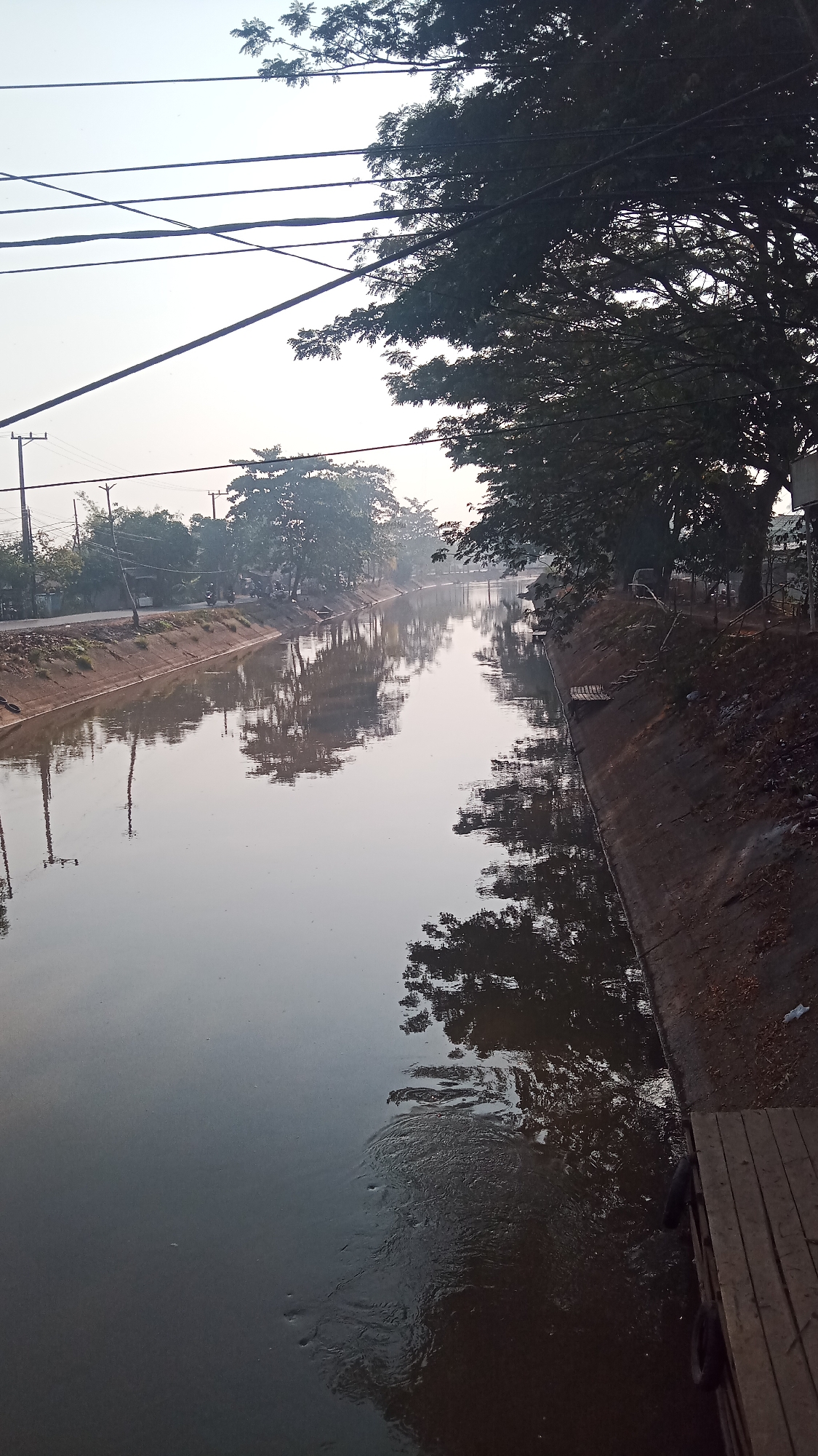 The image captures a canal running through Tanjung Rema, Indonesia, likely during the early morning or late afternoon given the soft lighting and diffused sky. The canal is lined with concrete banks and features reflections of trees and utility poles in its calm waters. A dirt road and vegetation run along the left bank, with several utility poles and wires visible. The right bank has a slope covered in dirt and debris, with a wooden structure and tires at the water's edge. The overall atmosphere is tranquil, with a sense of everyday life in a semi-urban, waterside environment.