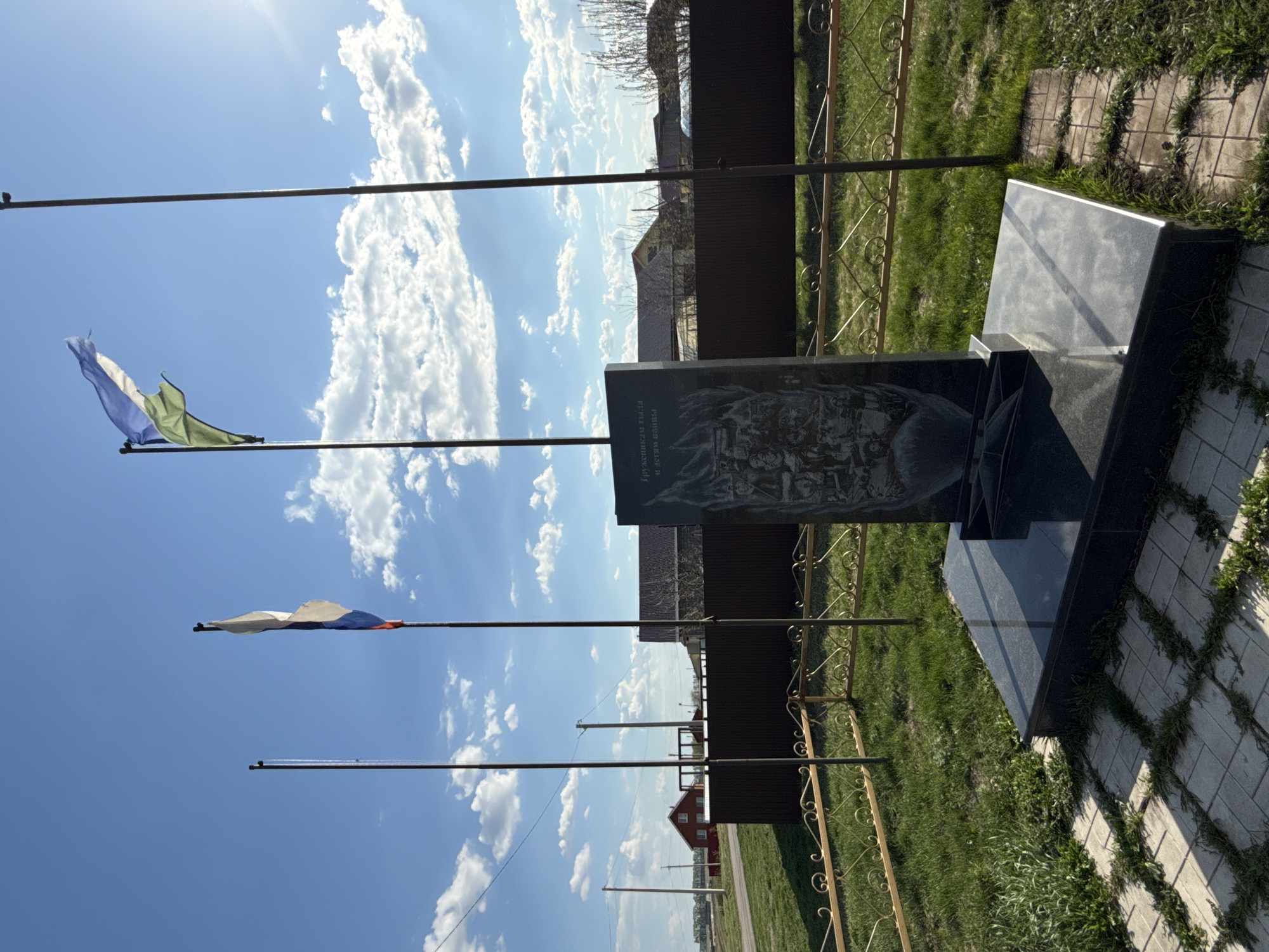 The image depicts a memorial in Takarlikovo, Russia. The memorial is made of dark stone and features an engraved image on its front panel. Behind the memorial is a brown fence, green grass, and some buildings in the distance. In the foreground, there are three flag poles. One flag is blue, white, and green, while another is white, blue, and red, resembling the Russian flag. The sky is blue with scattered white clouds.