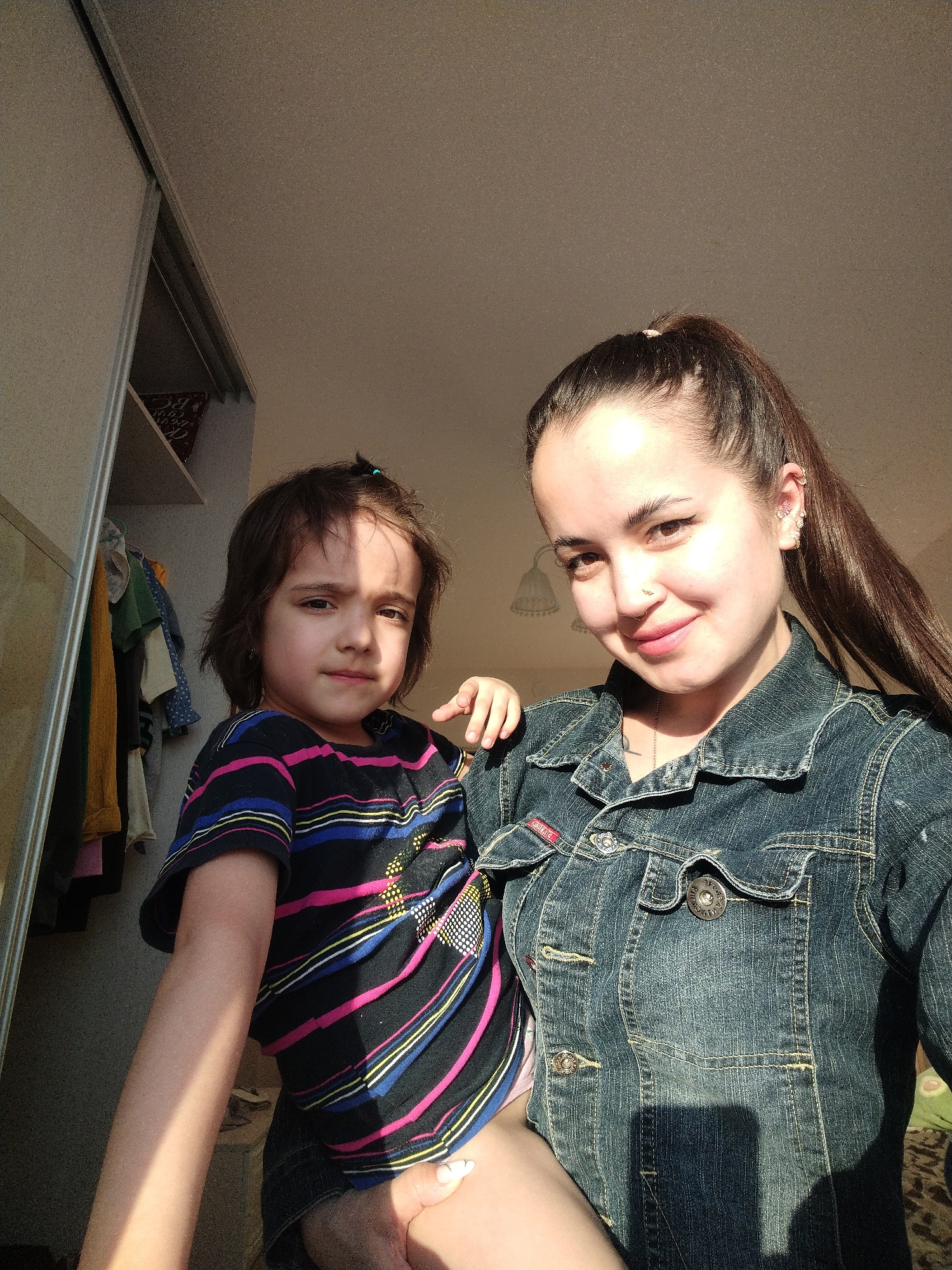 A woman wearing a denim jacket is holding a young girl with a striped shirt in what appears to be an indoor location. The scene includes a closet visible in the background, and it seems to be a well-lit area. The specific location within the city of Noviy, Russia cannot be determined from this image.
