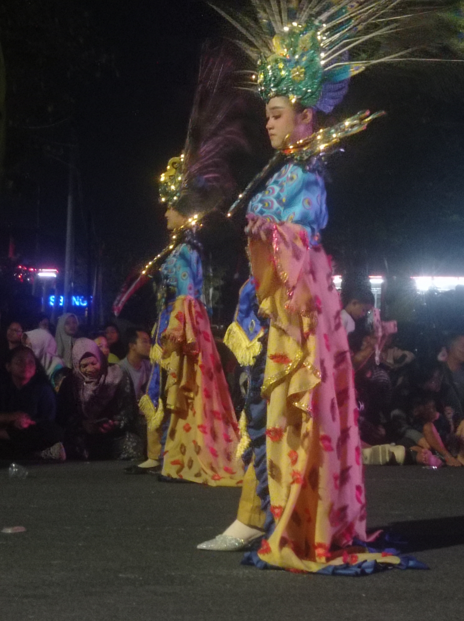 In Pandean, Indonesia, a nighttime cultural event is underway. Two individuals, likely performers, are the central focus. The person on the right is dressed in an elaborate costume featuring a tall, feathered headdress adorned with green and blue accents and small lights. Their garment consists of a blue, peacock-feather-patterned top and a flowing pink and yellow skirt with red floral designs. Golden, lighted embellishments are visible around the neck and shoulders. The second performer, partially visible on the left, wears a similar blue and yellow costume with a pink overlay, and a feathered headdress.
A crowd of onlookers is gathered behind the performers, many seated on the ground. The background is dark, suggesting it is night, with some blurry lights and structures visible, including a sign with blue neon lettering that reads "S NG". The ground appears to be asphalt. The overall atmosphere is one of celebration and cultural display.